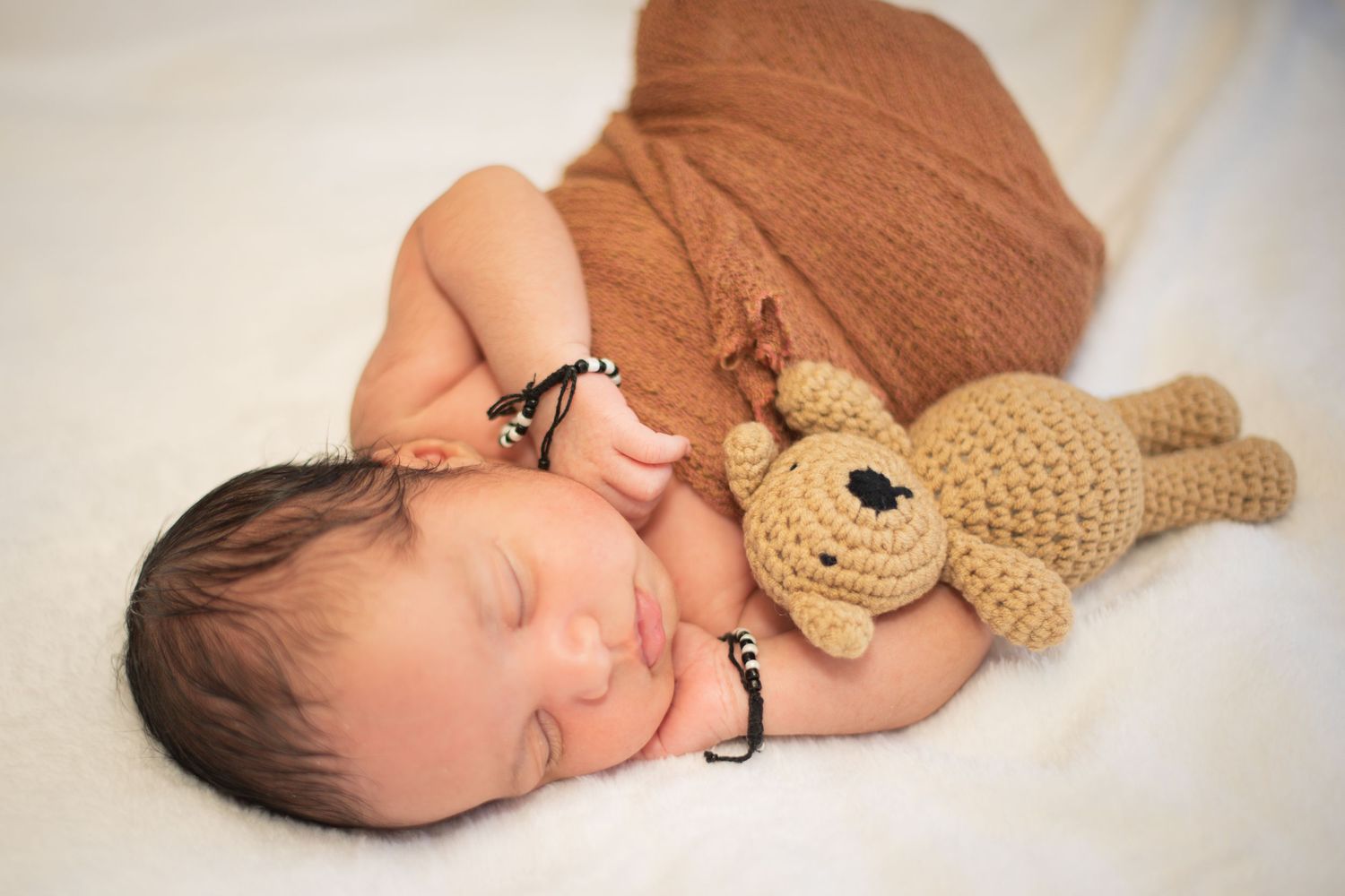 Sleeping newborn baby wrapped in brown knit blanket with crocheted teddy bear on white background.