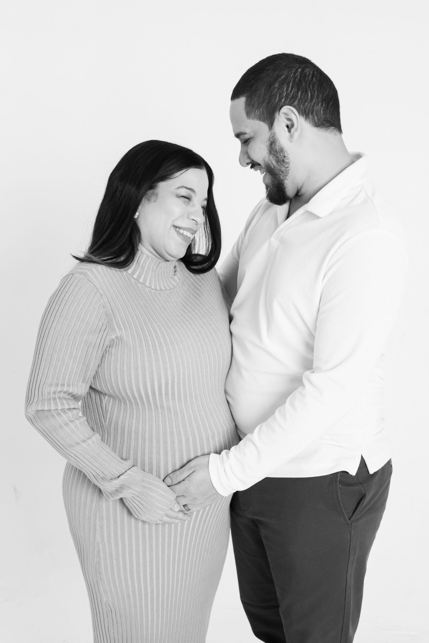 Black and white photo of a smiling pregnant couple tenderly embracing, the man gently touching the woman's baby bump.