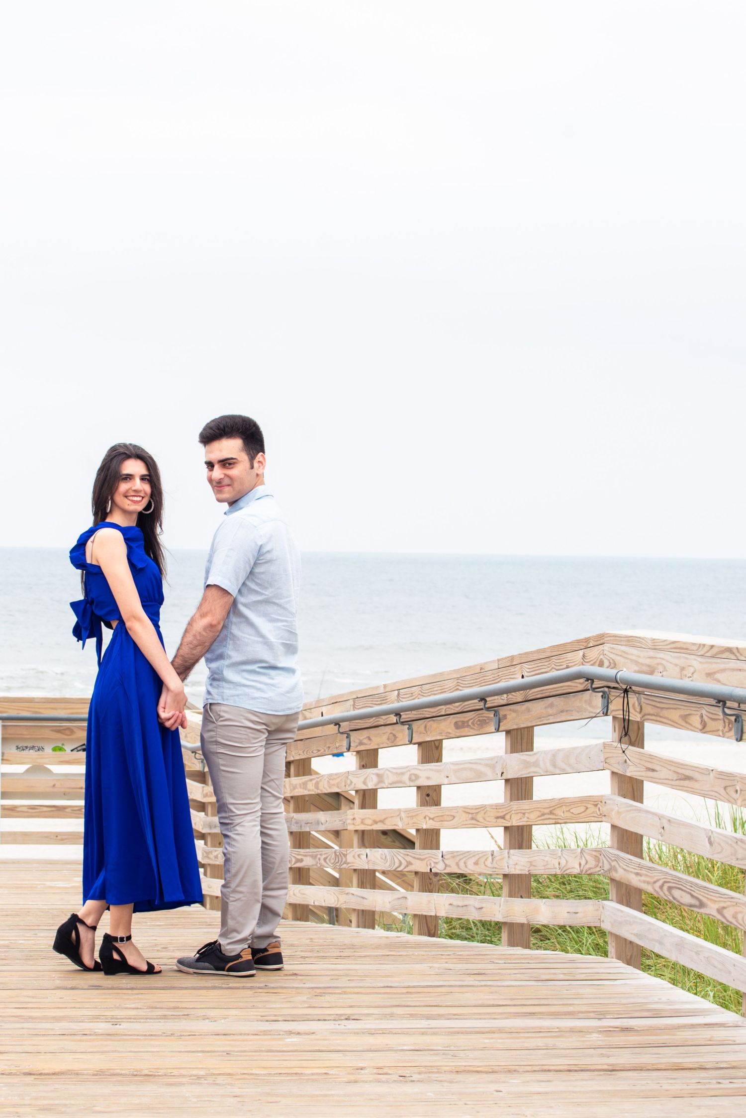 Couple holding hands on a wooden boardwalk by the ocean, woman in blue dress, man in light blue shirt and gray pants.
