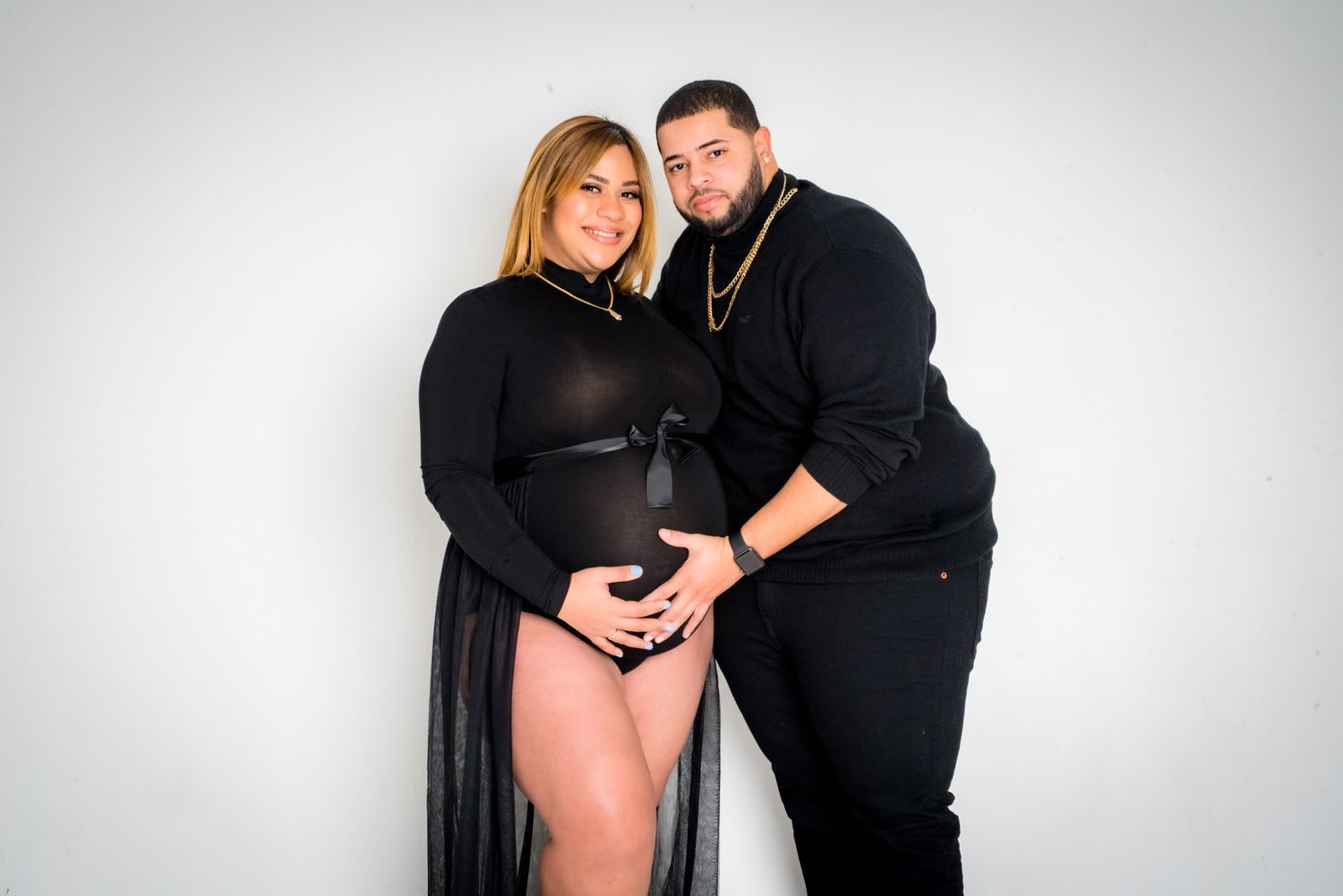 Couple in matching black outfits posing for a maternity photo shoot against a white background.