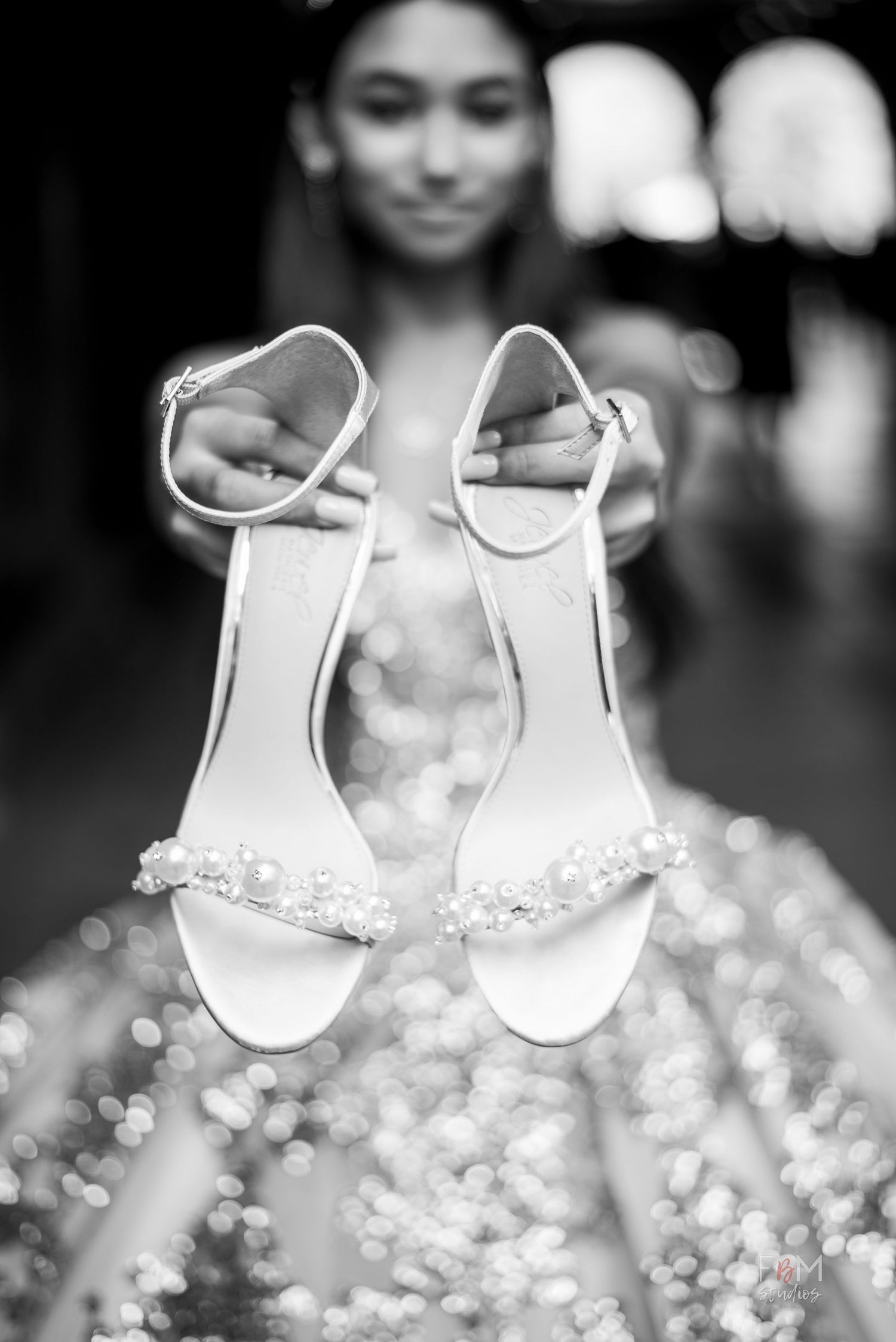 Black and white photo of a bride holding pearl-embellished heeled sandals with bokeh sparkle dress background.