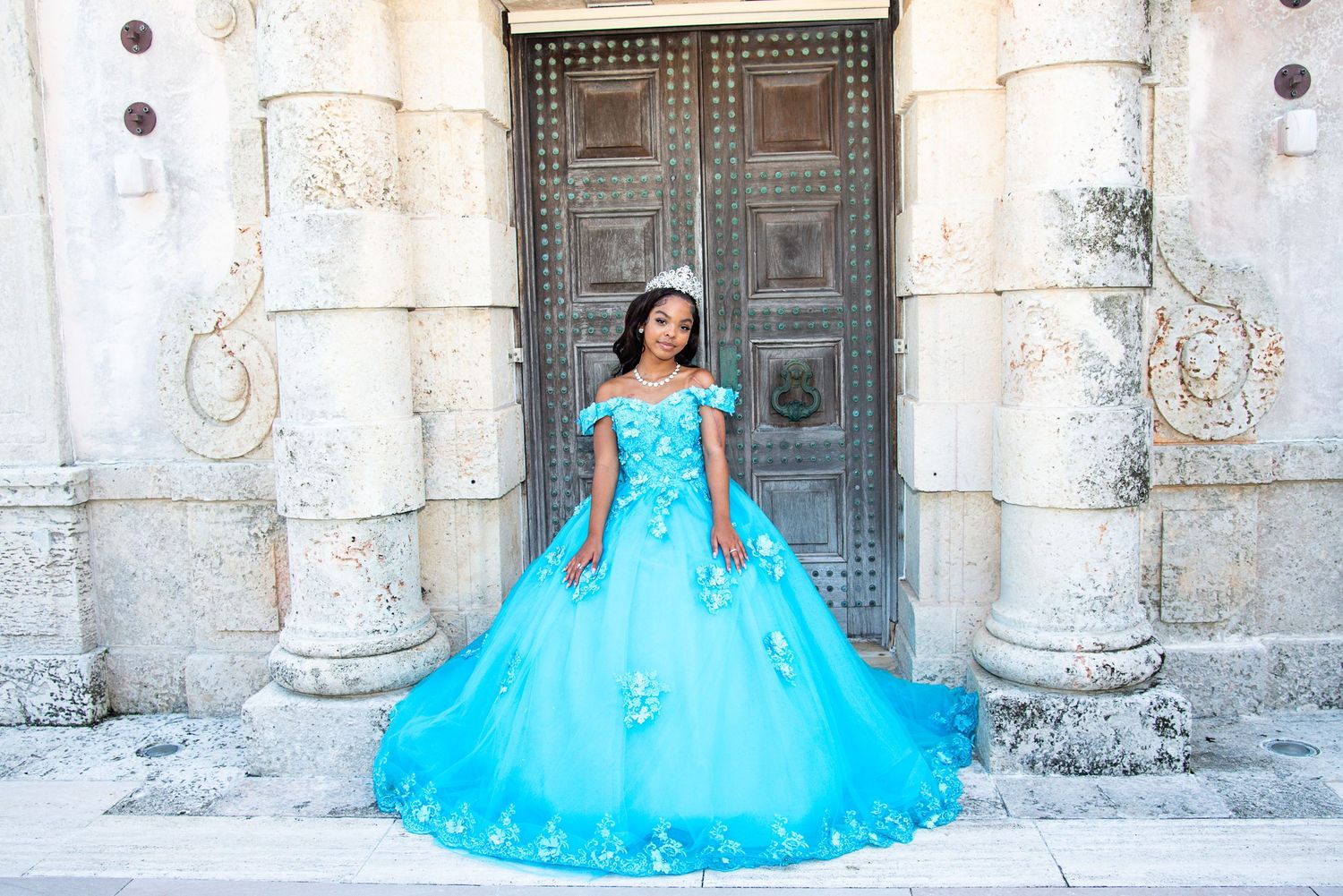 Young woman in a stunning turquoise ball gown and tiara poses elegantly before ornate doors for her Quinceañera.
