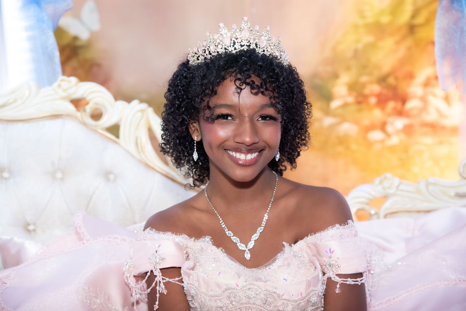 Young girl in white lace gown and tiara smiles at quinceañera celebration with pink floral backdrop.