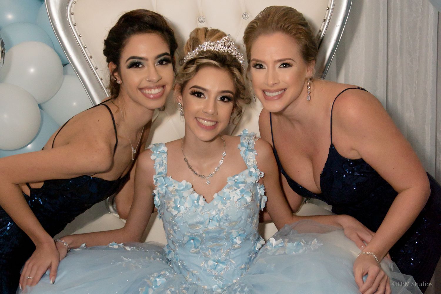 Three women pose together at a quinceañera celebration, with the birthday girl in a light blue ball gown and tiara.