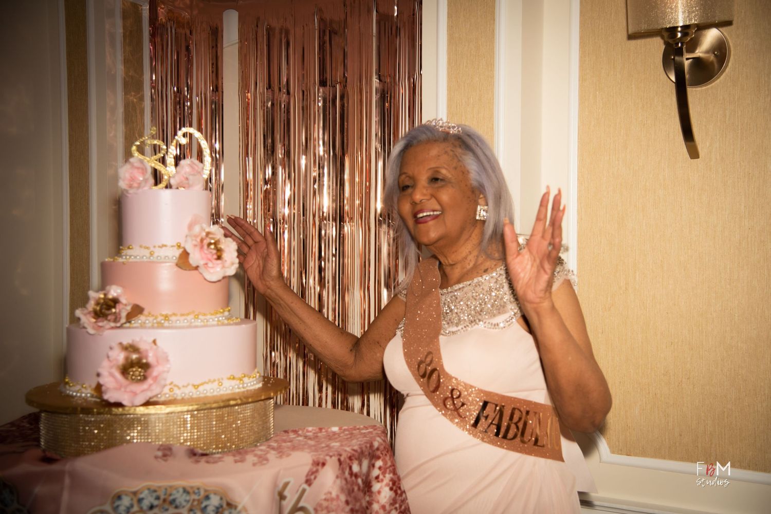 A celebrant in a white dress gestures joyfully next to an elegant pink tiered cake with floral decorations by FBM Studios in Fort Lauderdale