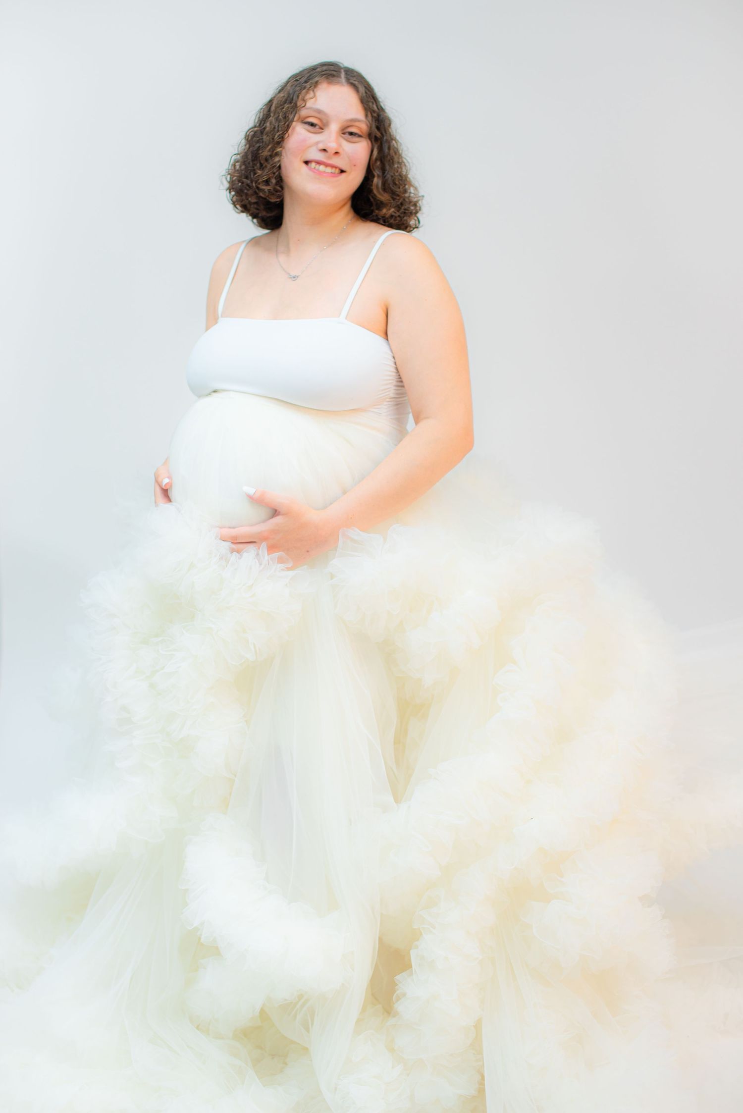 Smiling pregnant woman in white tulle maternity gown cradling baby bump against light background.