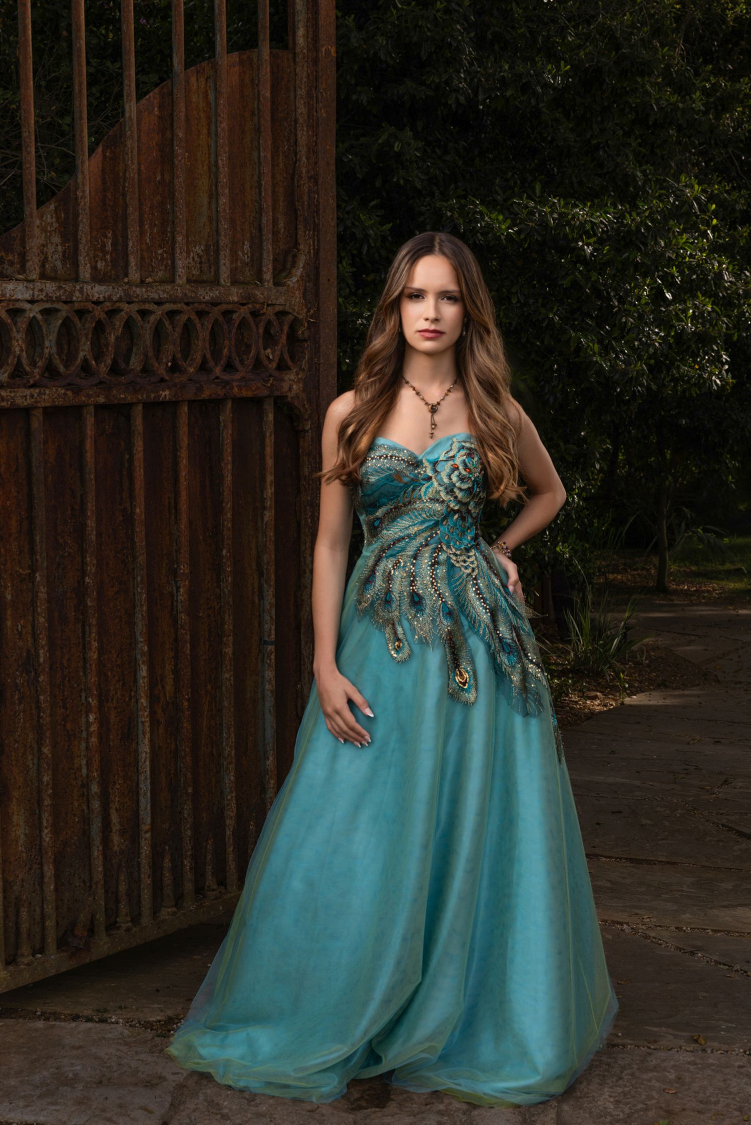 Young woman in elegant teal strapless ball gown with peacock embroidery poses near rustic iron gate outdoors.