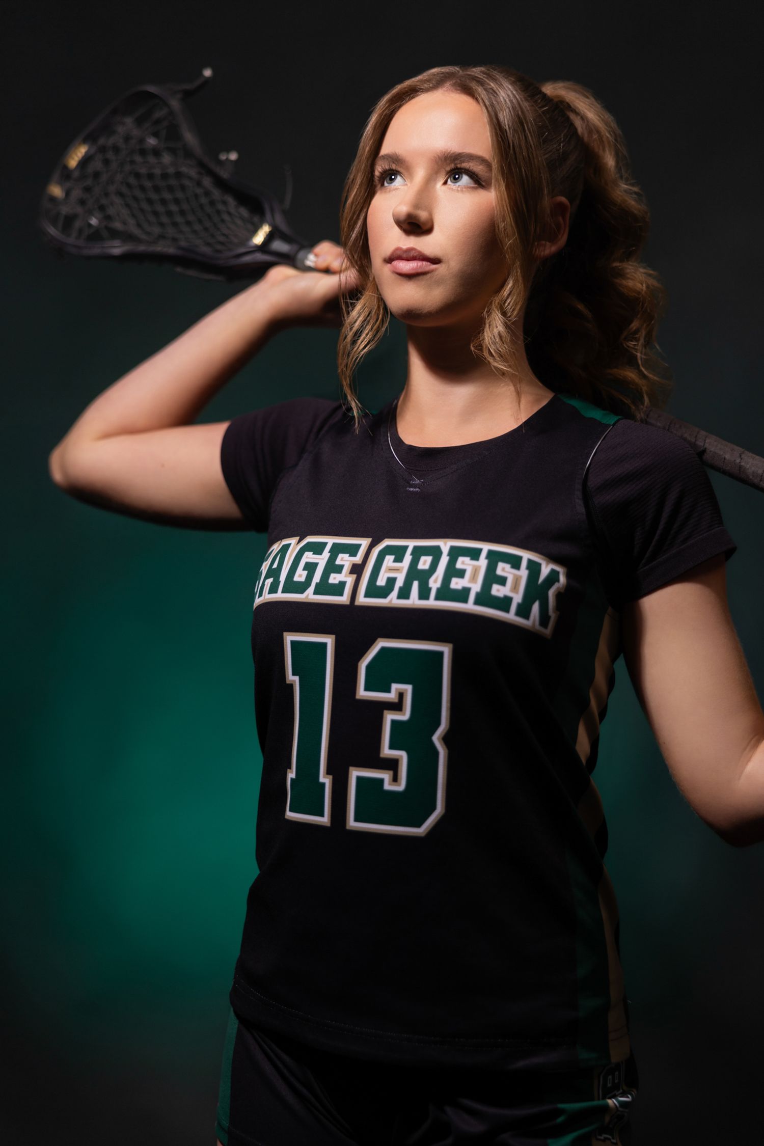 Teen girl in Sage Creek #13 lacrosse jersey holding stick over shoulder against dark green background.