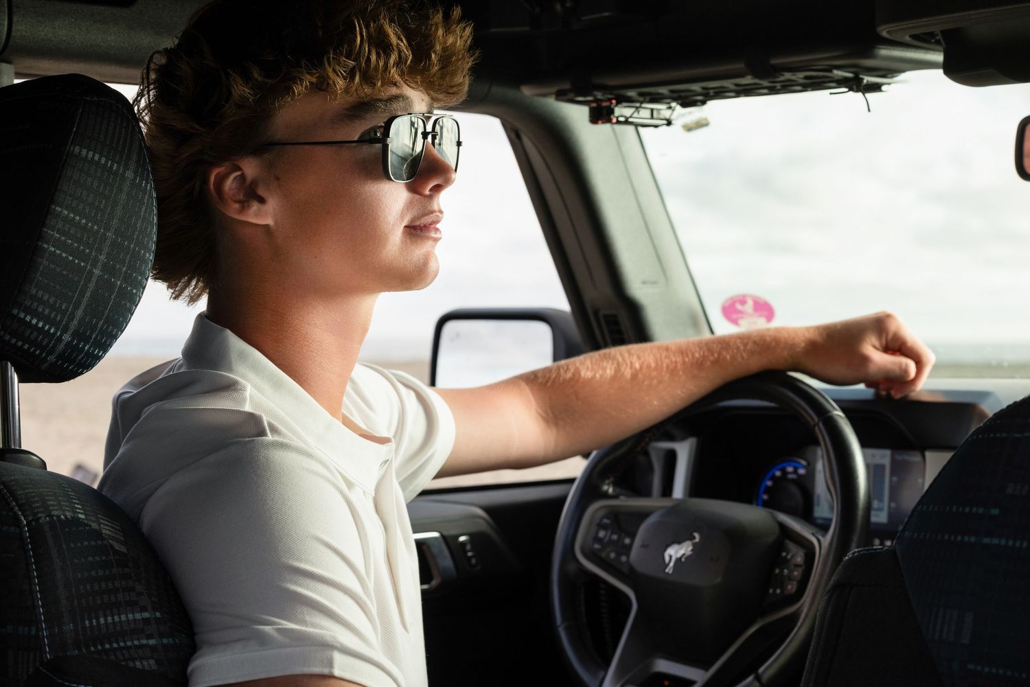 Young man with sunglasses driving a Ford vehicle, arm resting on steering wheel, casual white shirt.