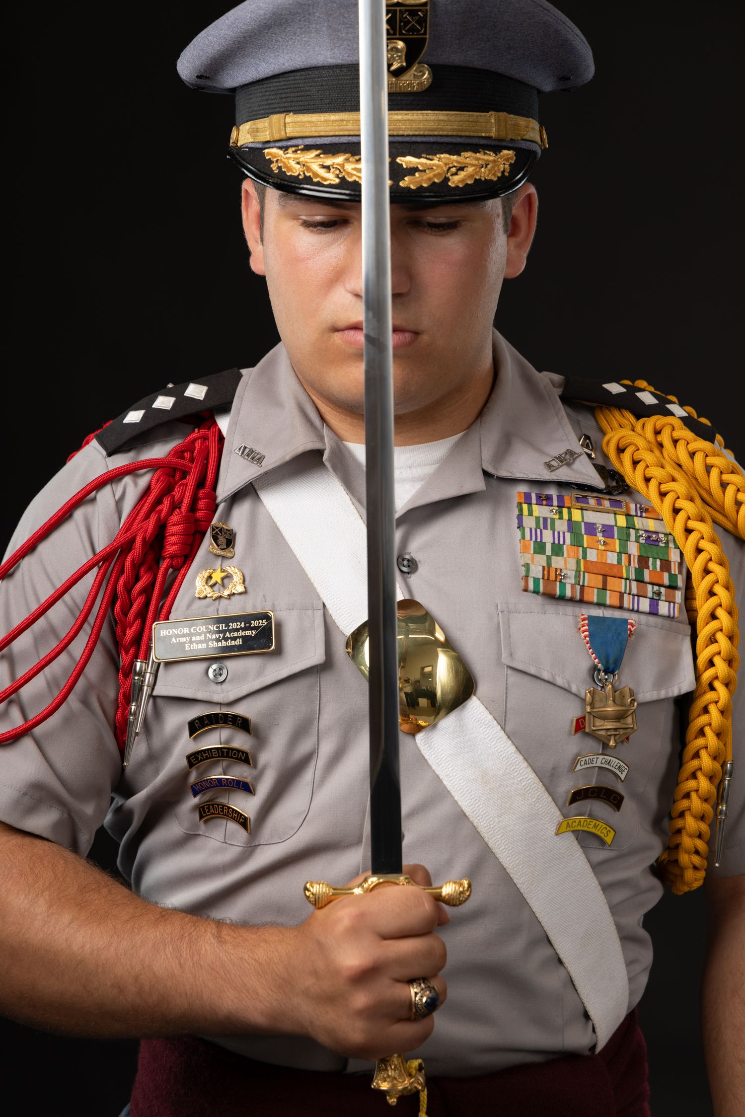 Military cadet in dress uniform holding a ceremonial sword, wearing medals and colorful shoulder cords.