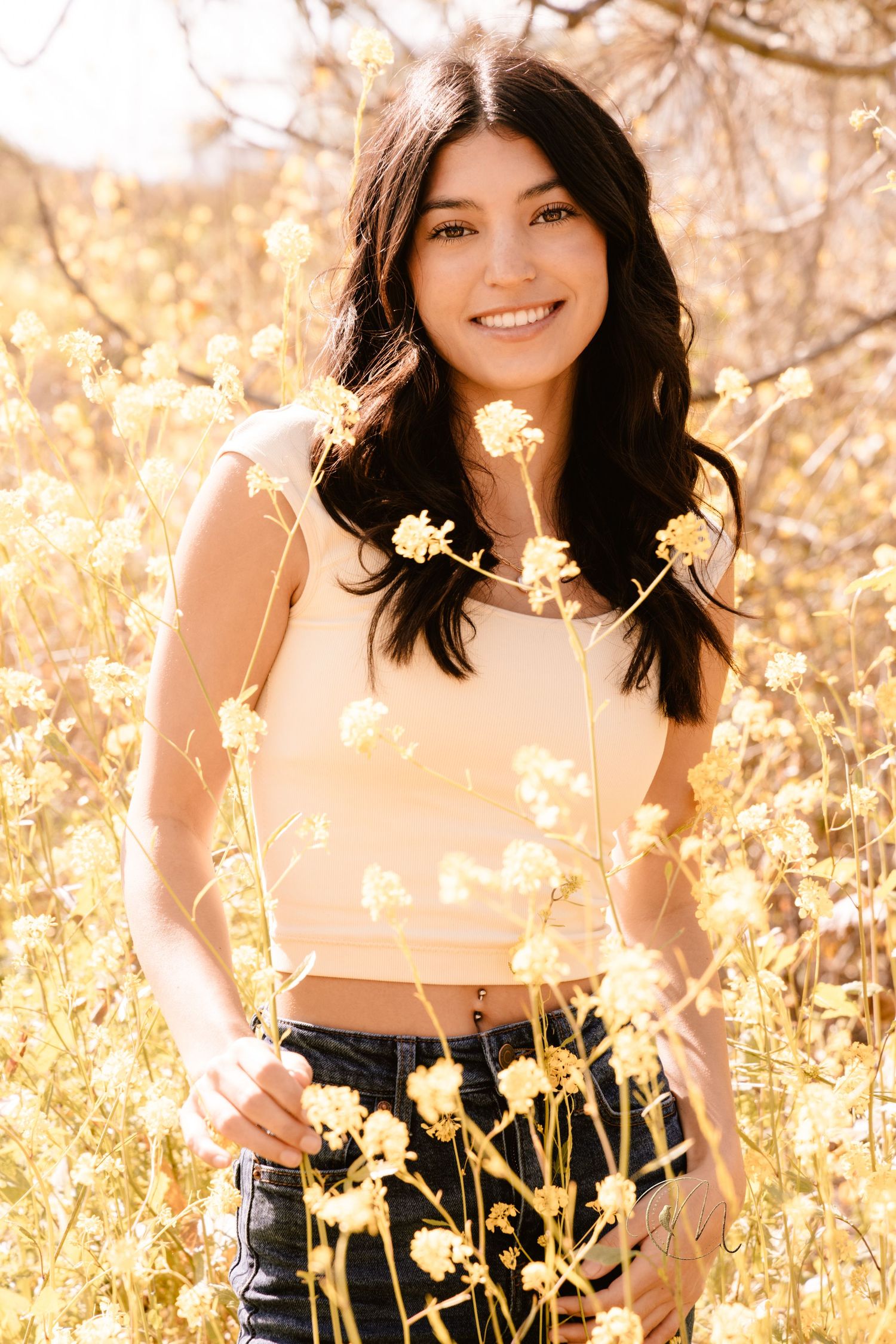 Smiling young woman with dark hair in a crop top and jeans standing among yellow wildflowers in a sunny field.