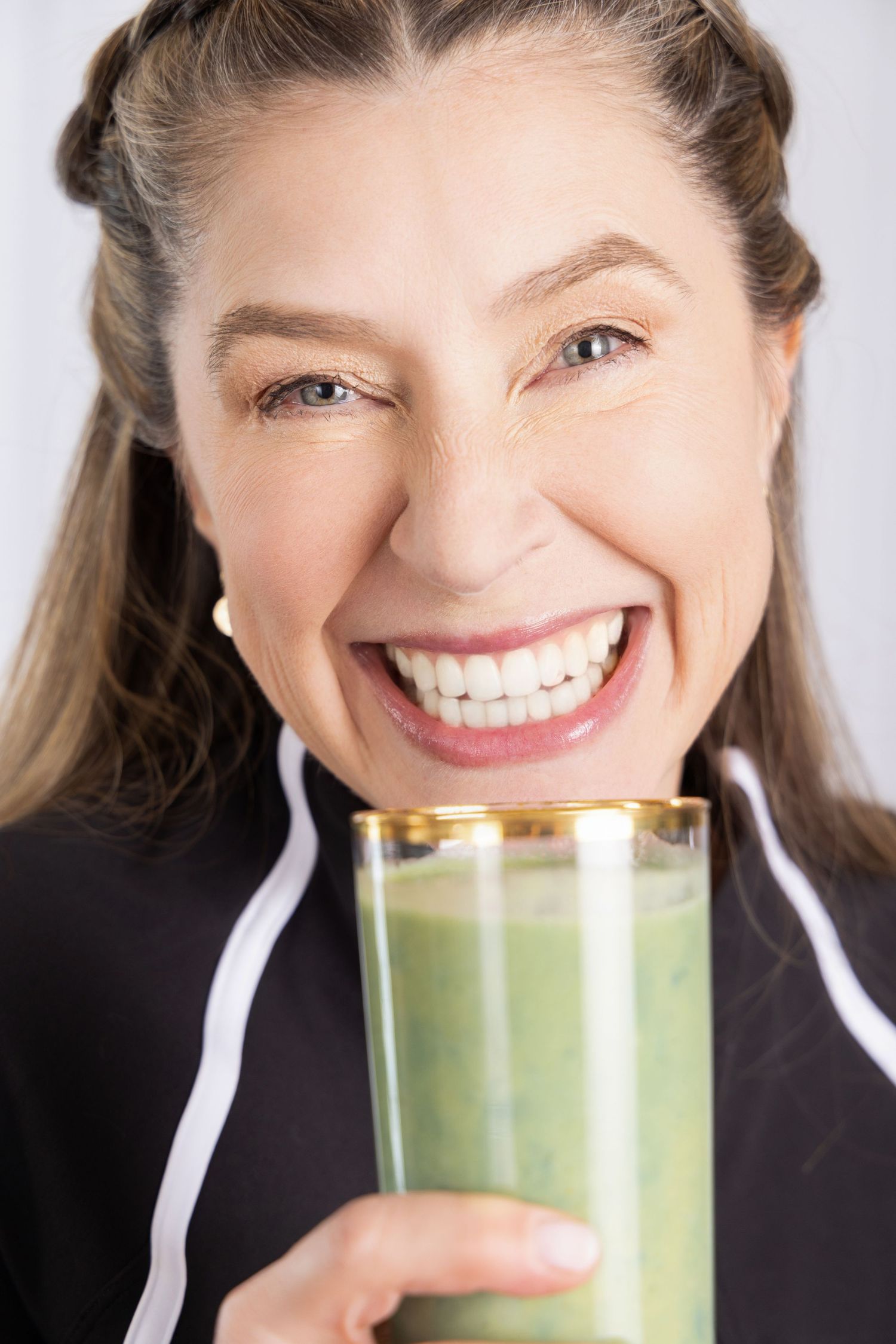 Smiling middle-aged woman holding a green smoothie in a glass, wearing a black athletic jacket against a white background.