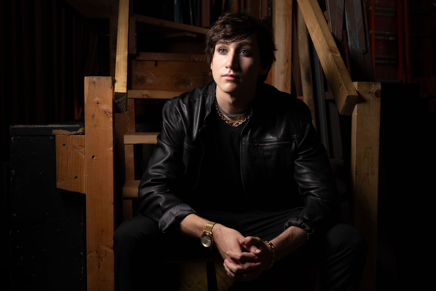 Young man in black leather jacket sits on wooden stairs in dramatic lighting.