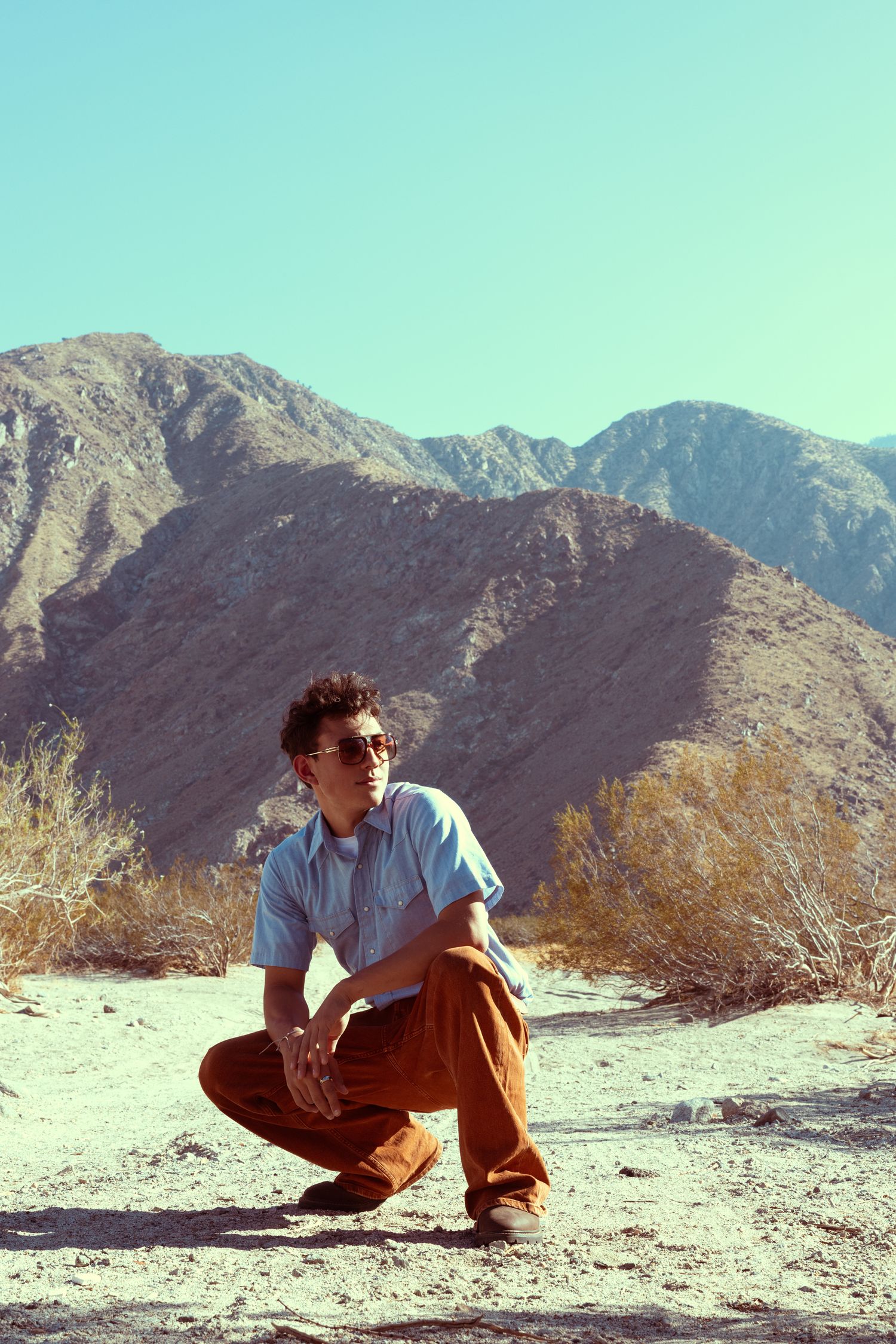 A young man in sunglasses and brown pants crouches in a desert landscape with mountains and clear blue sky behind him.