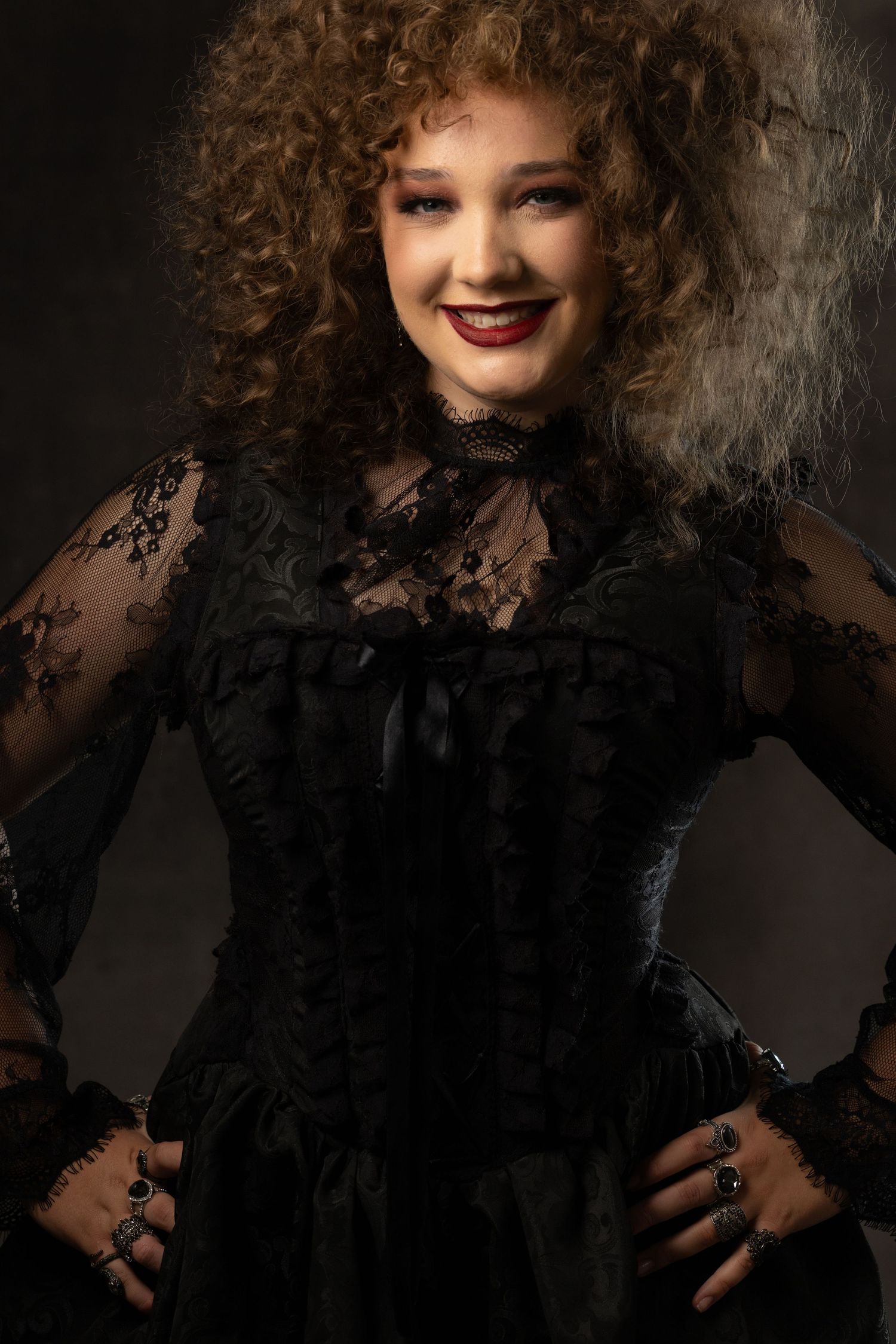 Smiling woman with curly hair wearing a black lace corset gothic dress with rings, posing with hands on hips.