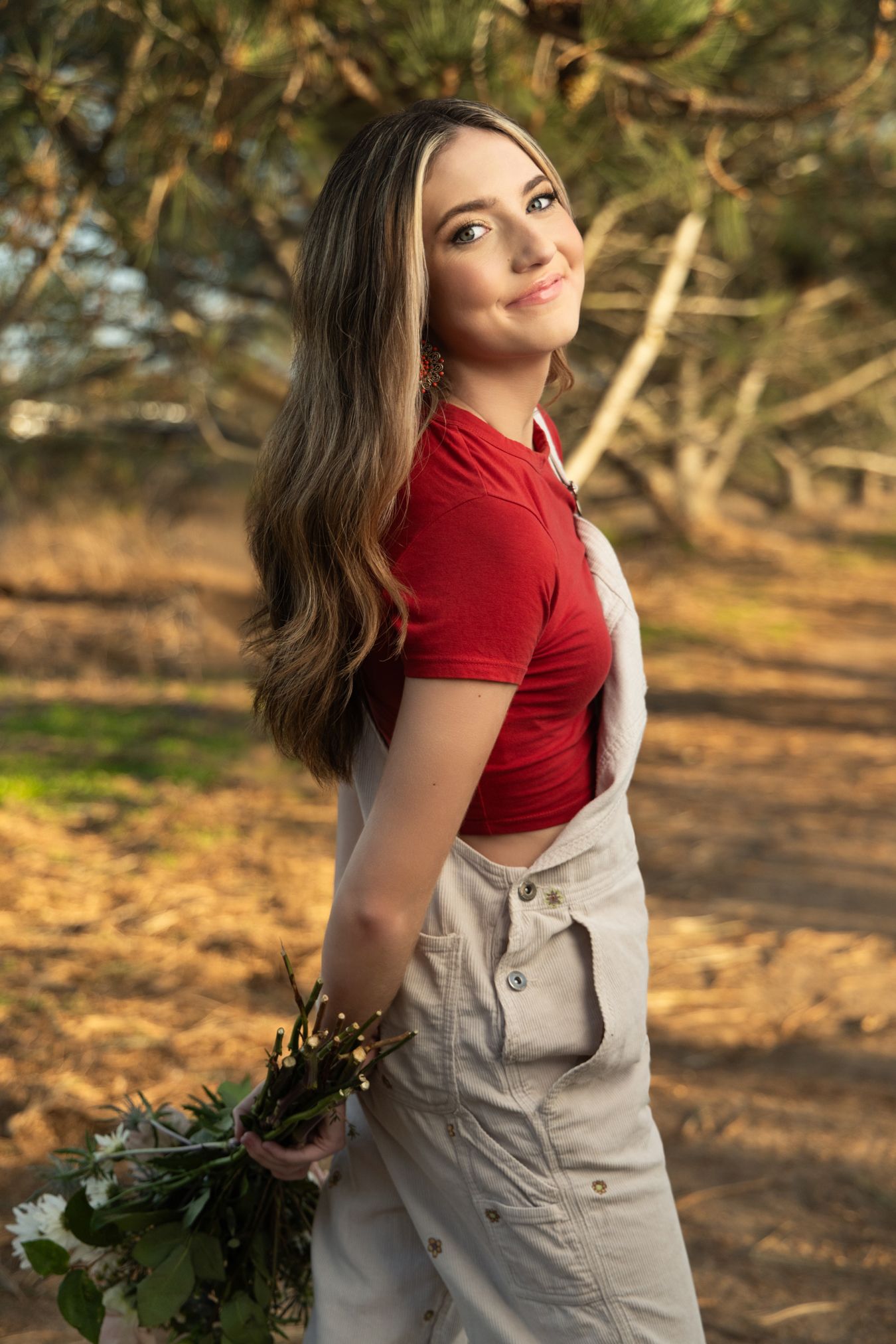 Smiling teenage girl in red top and overalls holding wildflowers in golden outdoor setting.