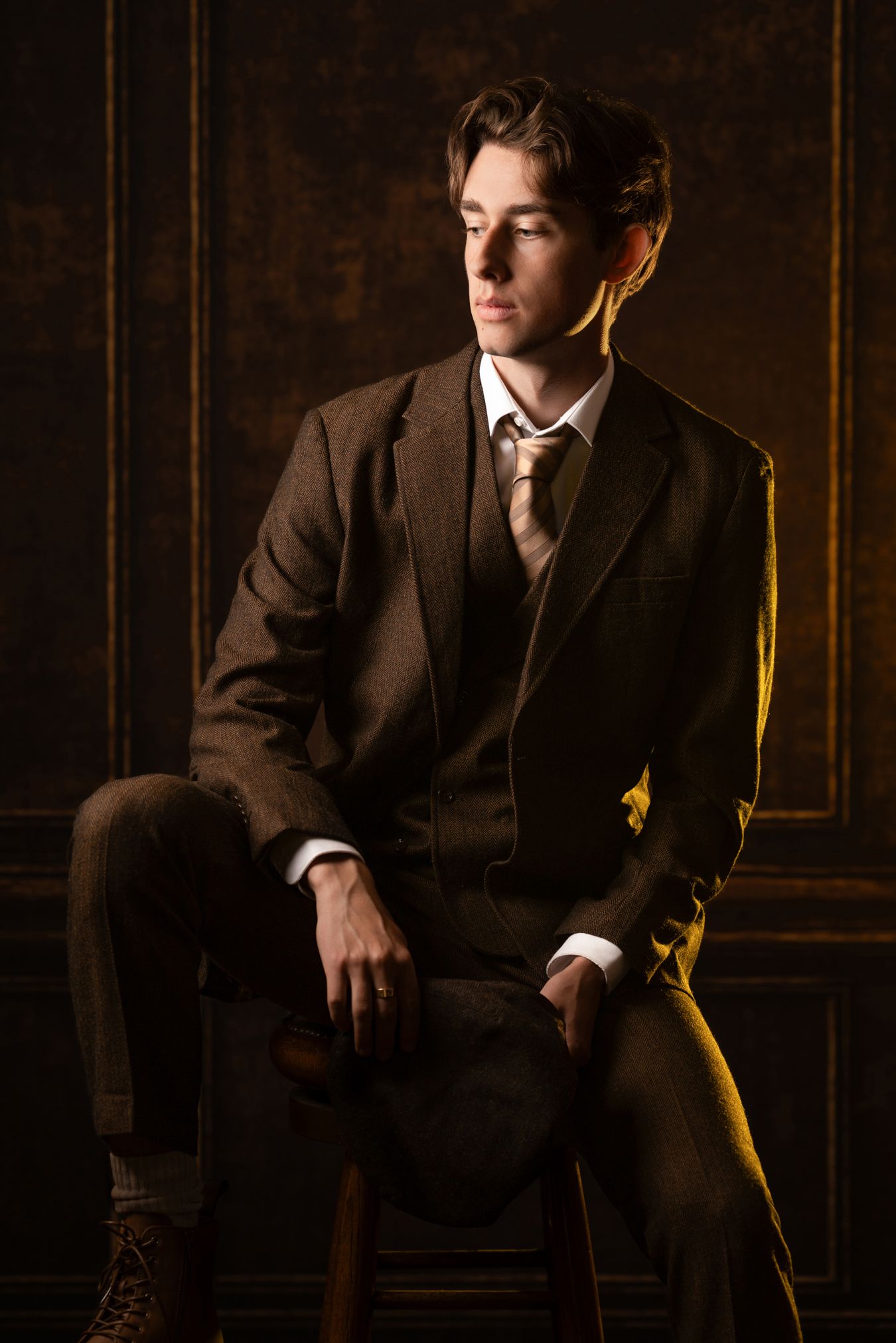 Elegant young man in brown tweed three-piece suit seated on stool with warm golden studio lighting.