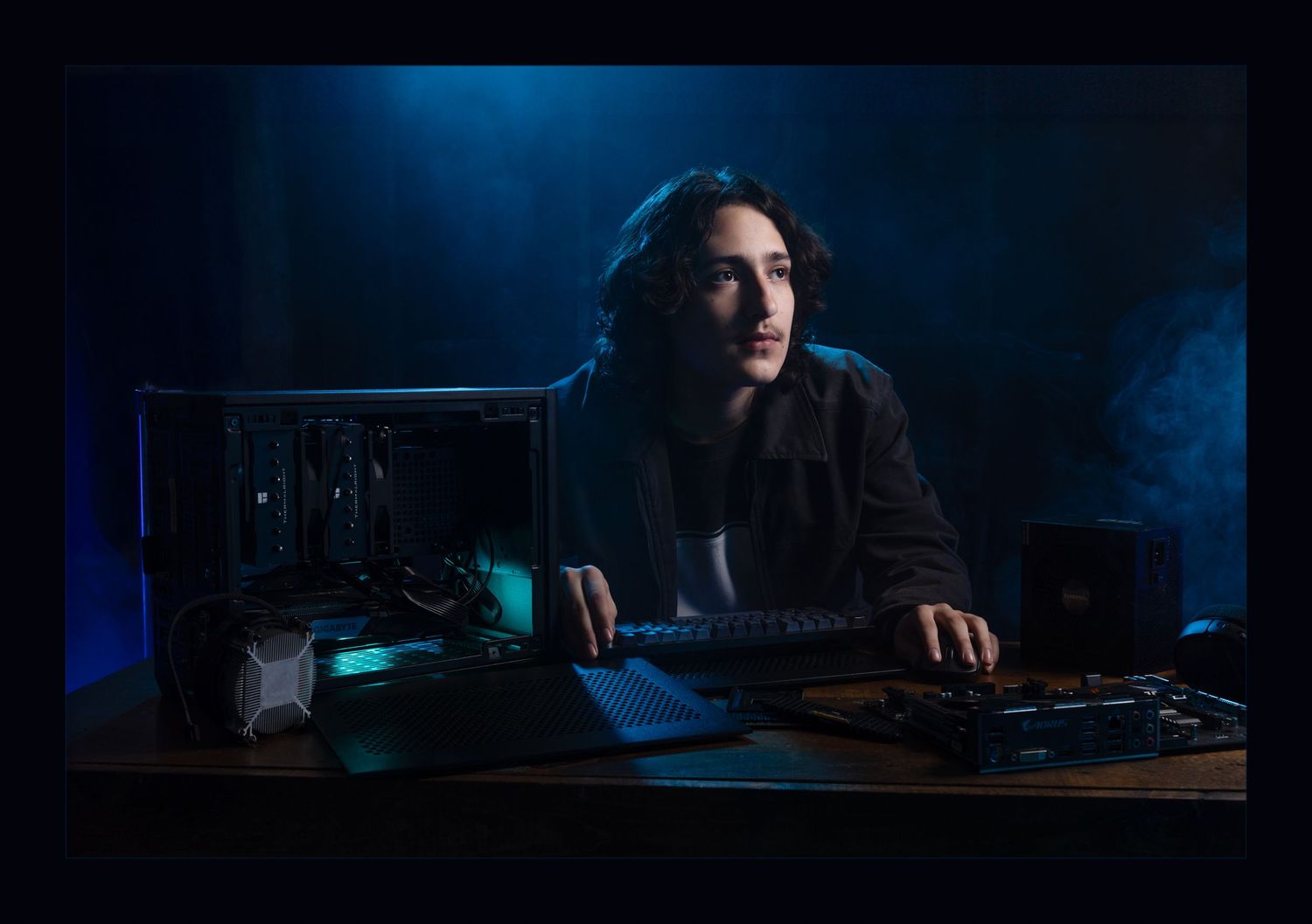 Young man sits at gaming PC setup with blue atmospheric lighting in dark room.