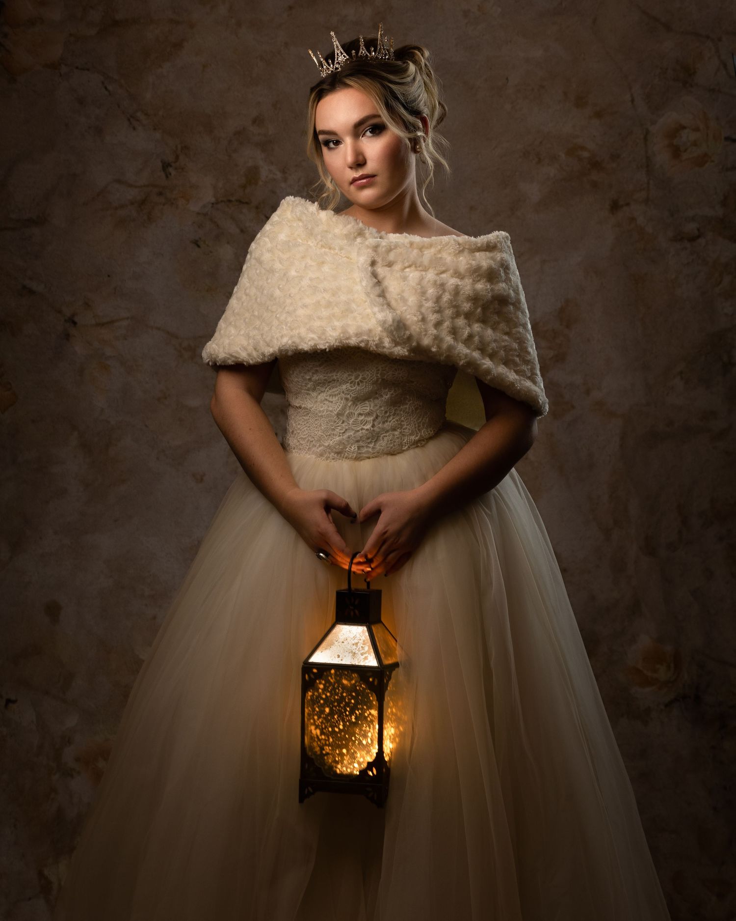 Young woman in a white tulle gown and fur shawl wearing a crown holds a glowing lantern in a portrait.