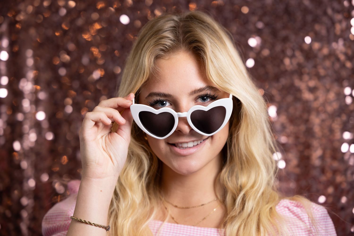 Smiling blonde woman wearing white heart-shaped sunglasses against a rose gold sequin backdrop.