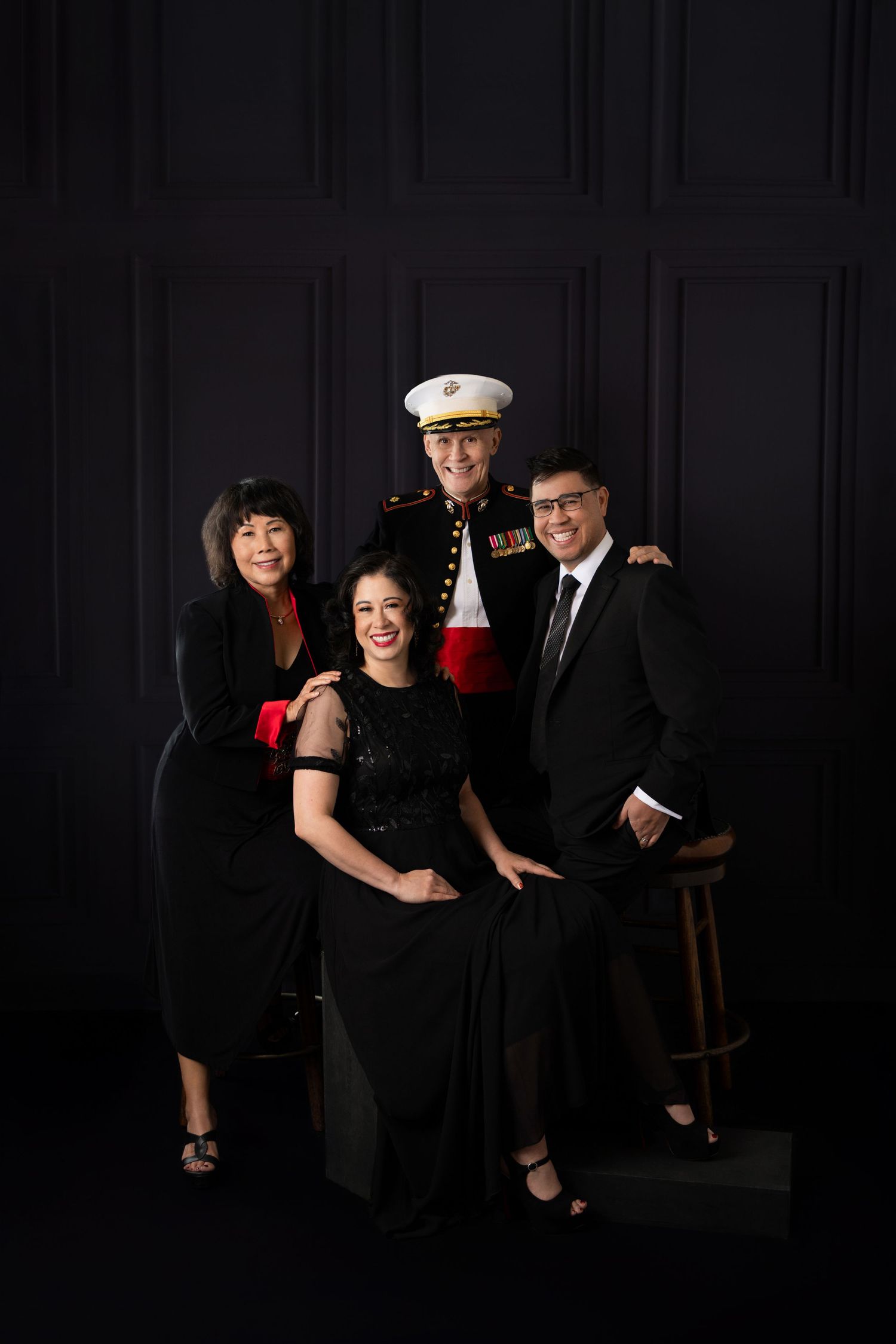 Family portrait with four people in formal attire, one in U.S. Marine dress uniform, posed against a dark background.
