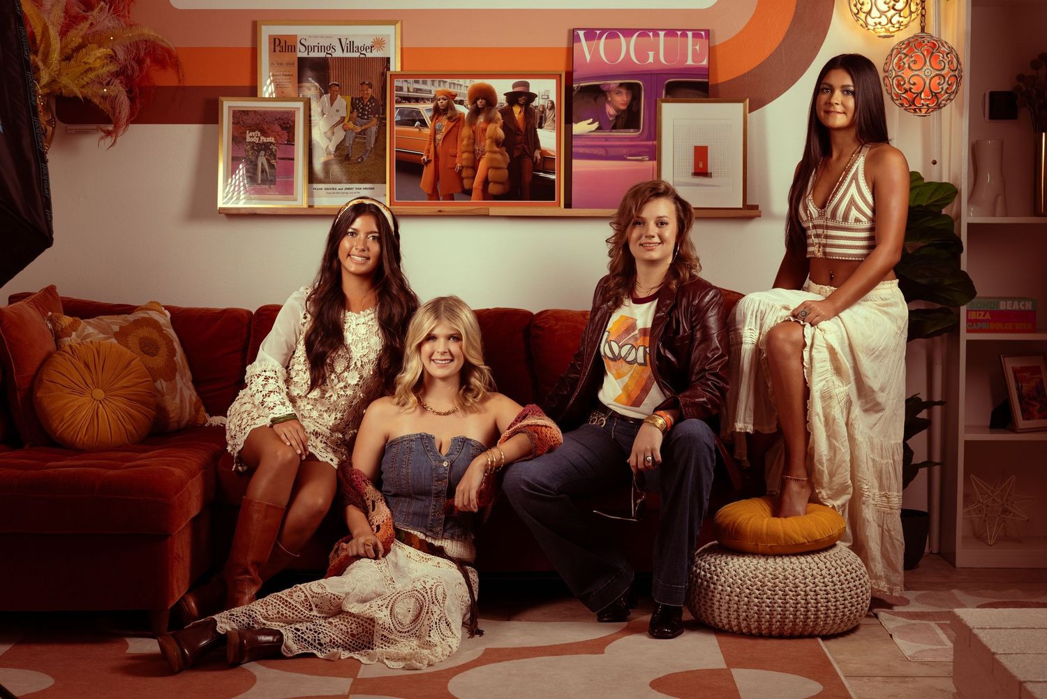 Four women pose in a retro 70s-inspired room with vintage posters, warm tones, and a cozy orange sofa.