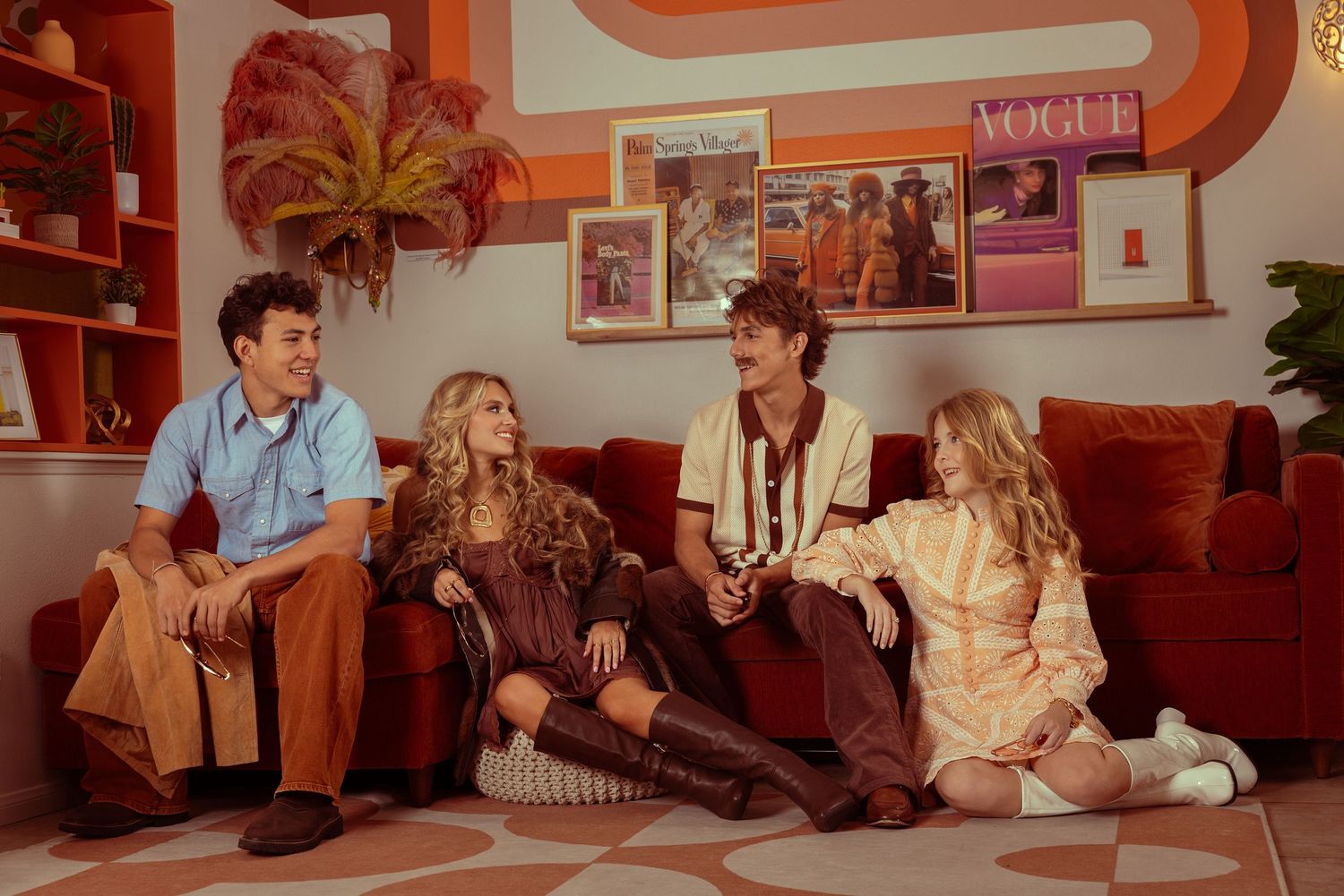 Four friends in 70s-inspired outfits relax on a red sofa in a retro-decorated room with warm tones.