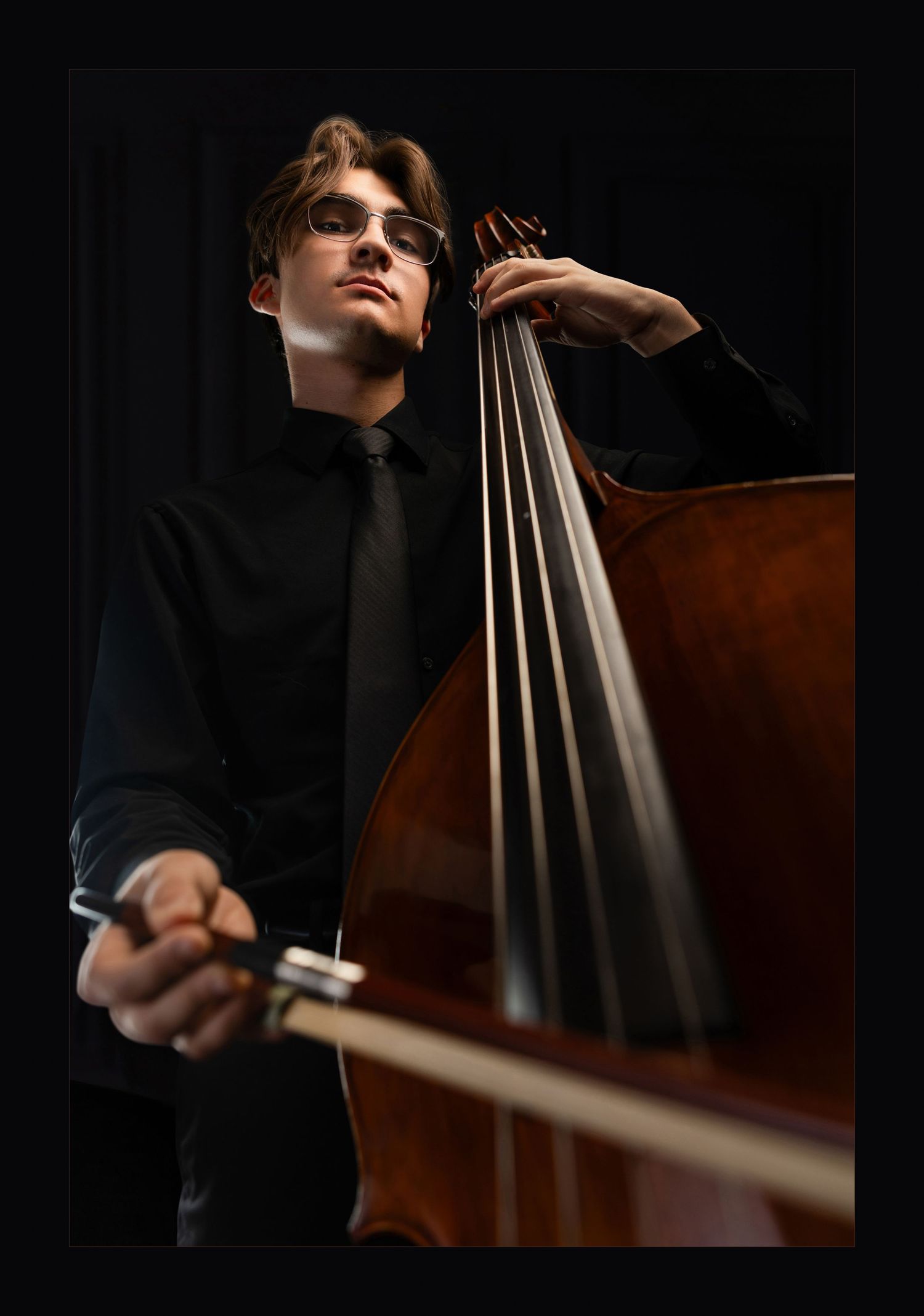 Young male musician in black attire playing double bass, holding bow, wearing glasses against dark background.