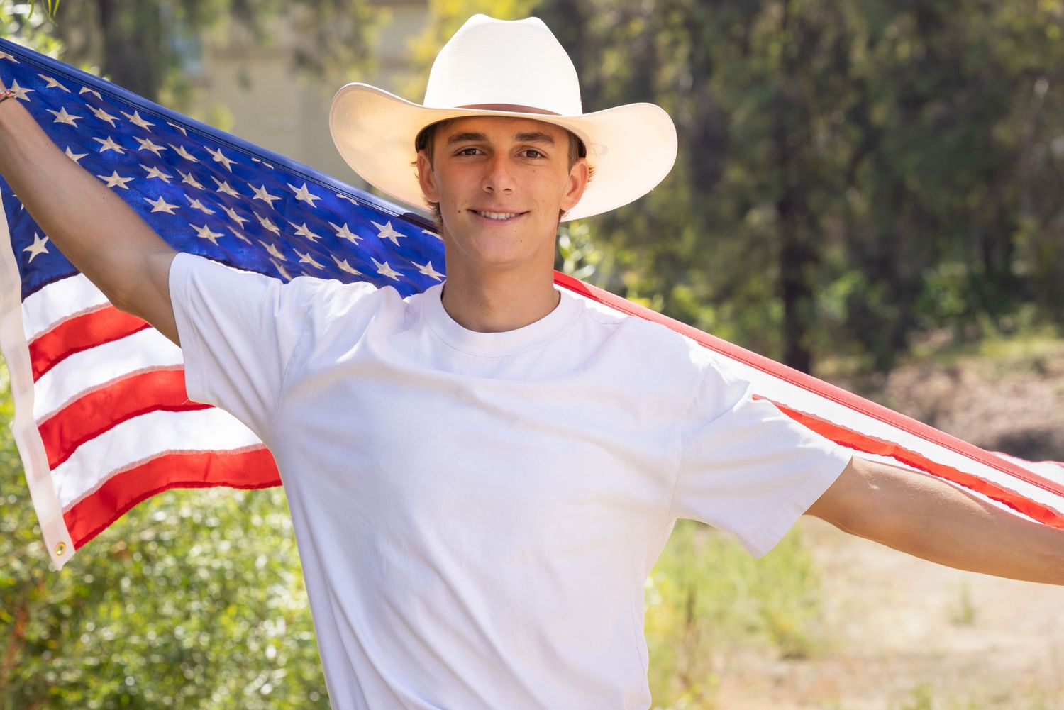 Young man in white cowboy hat proudly holding American flag outdoors, smiling in patriotic display.