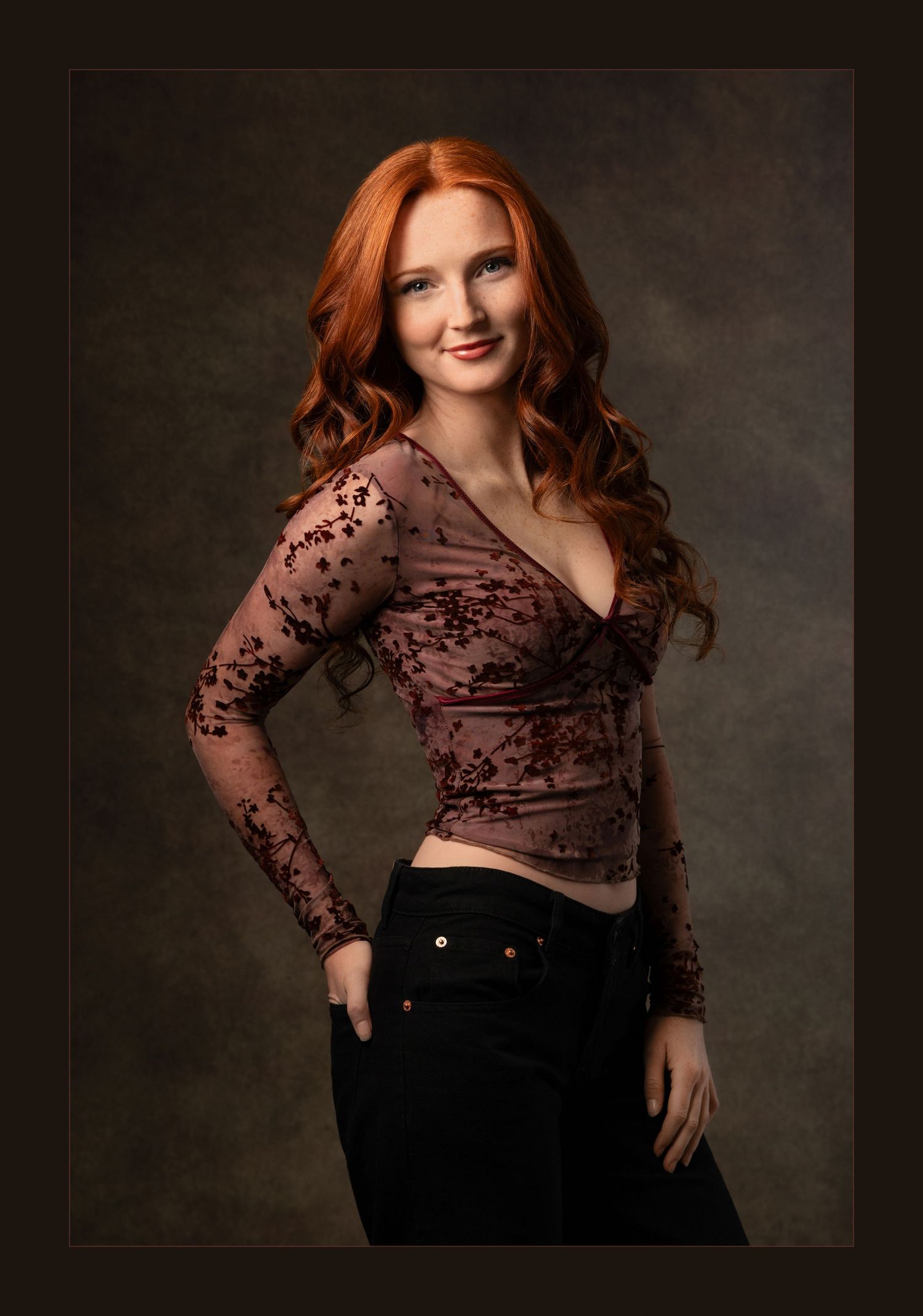 A young woman with long red hair poses confidently in a floral lace top and black jeans against a dark studio background.