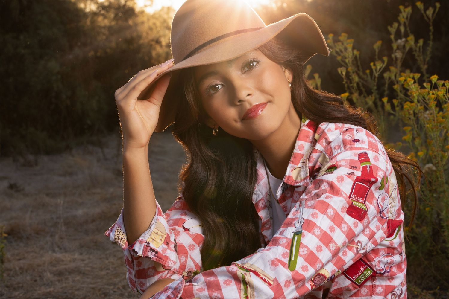 Young woman in a red gingham shirt and wide-brim hat smiling outdoors in golden sunlight.