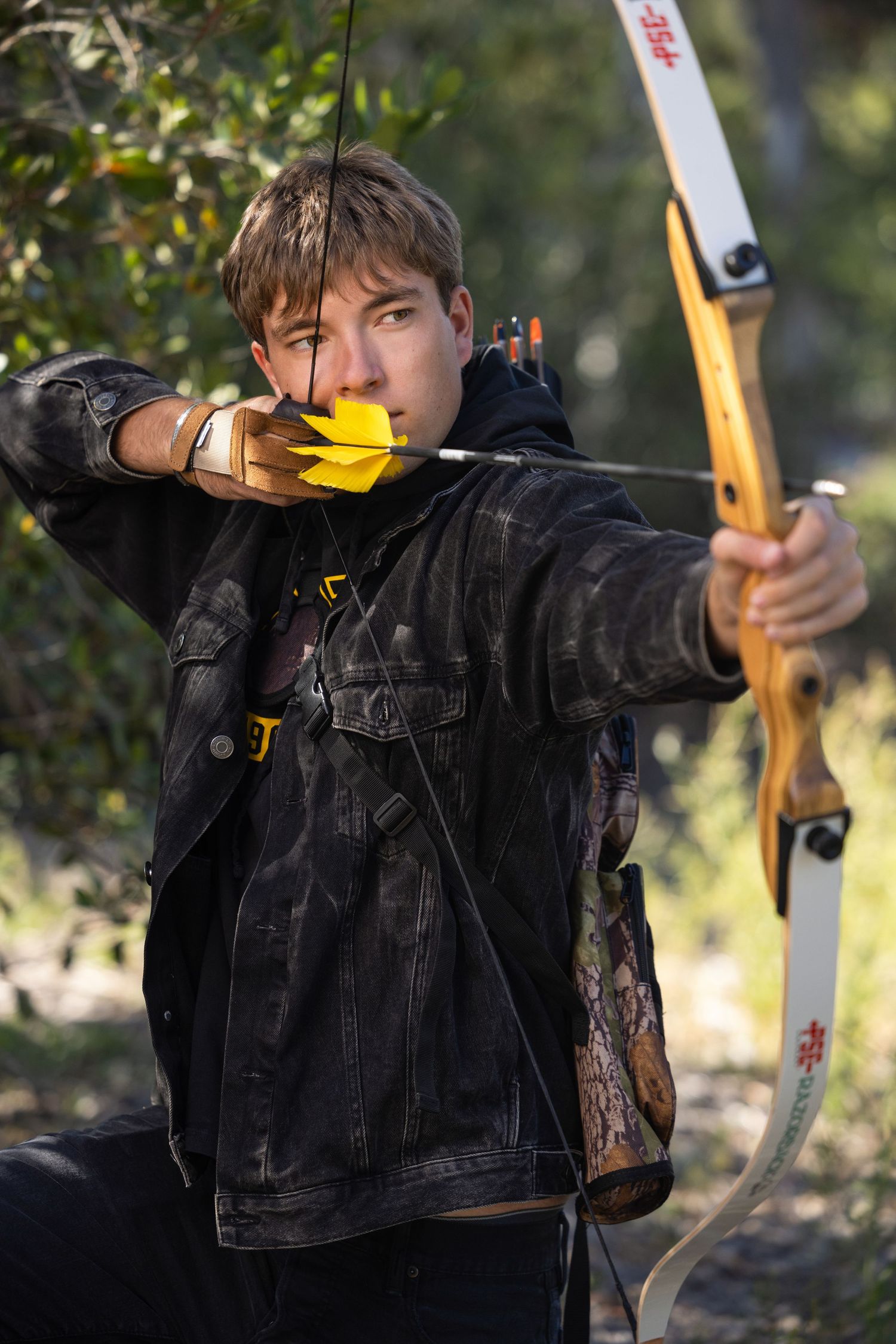 Young male archer in black jacket drawing a bow with yellow-fletched arrow in a forest setting.