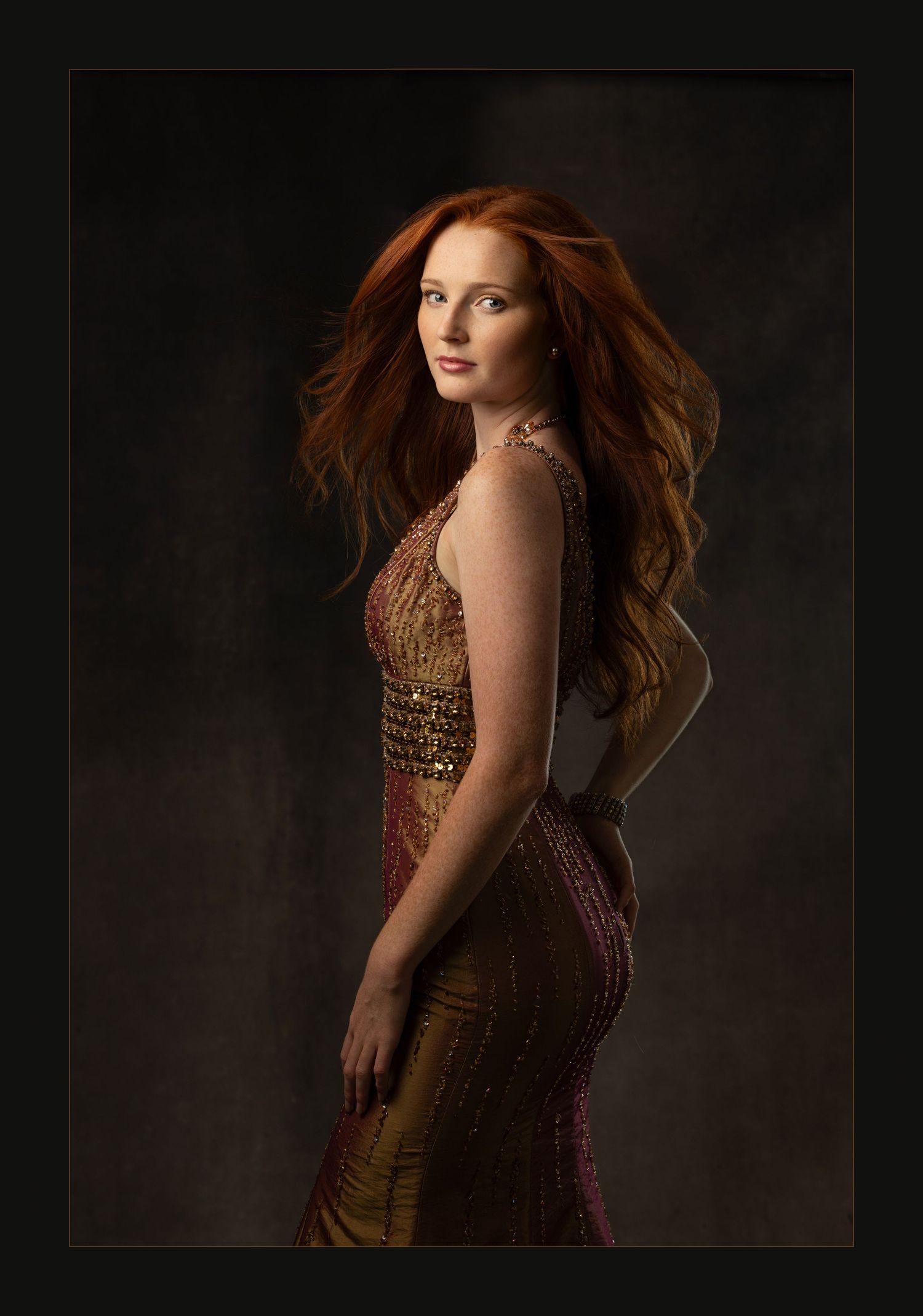 A stunning redhead woman poses in a gold sequined gown against a dark studio background.