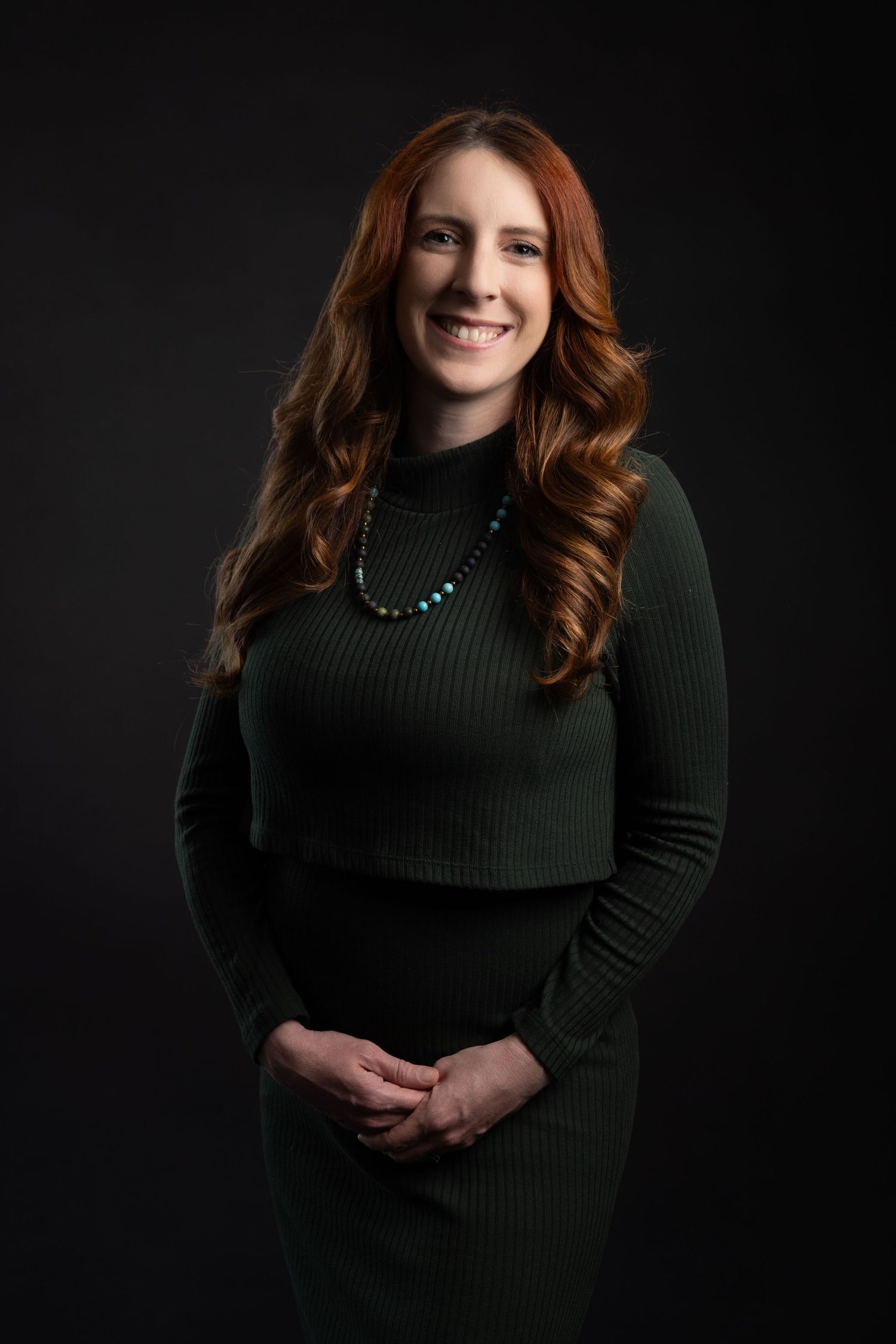 Professional woman with red hair in green ribbed dress and turquoise necklace smiling against dark background.