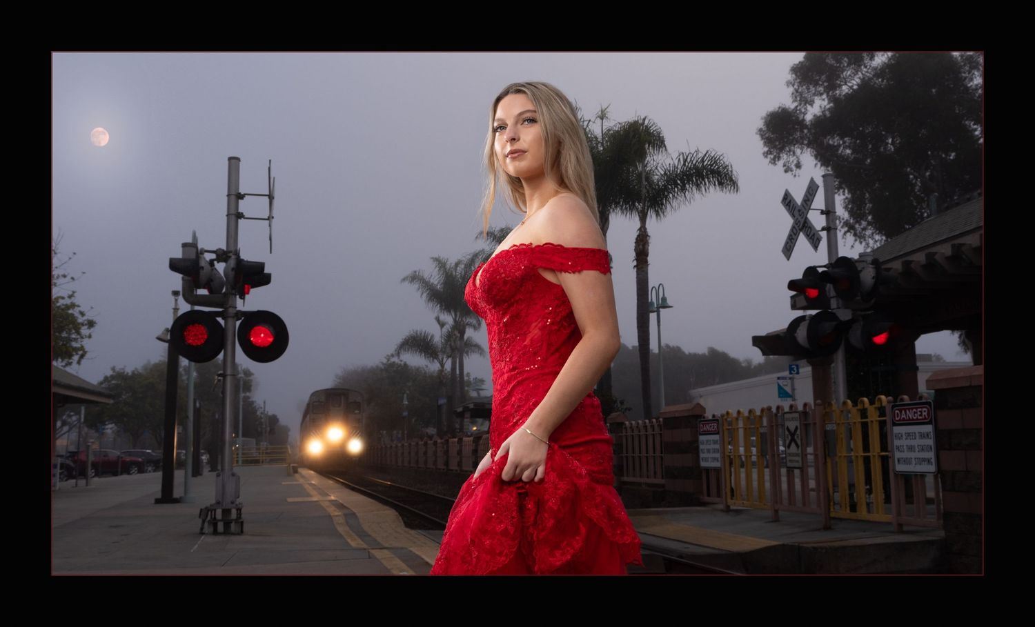 Blonde woman in red off-shoulder dress stands at railroad crossing in foggy evening.