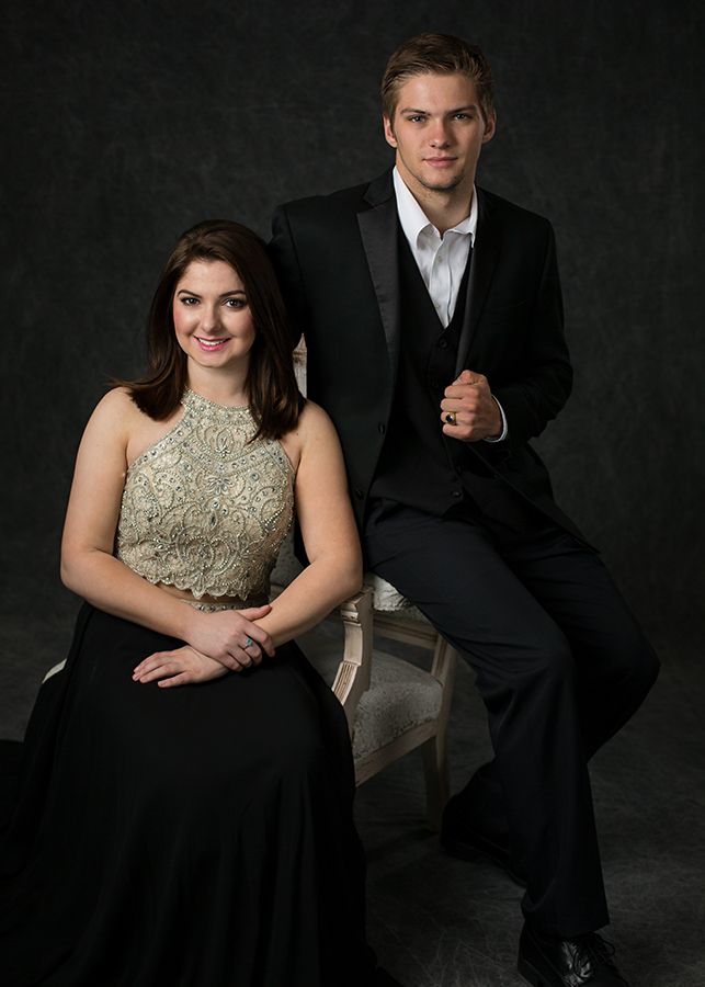 Young couple in formal attire posing for prom portrait, woman in gold gown seated, man in black tuxedo standing.