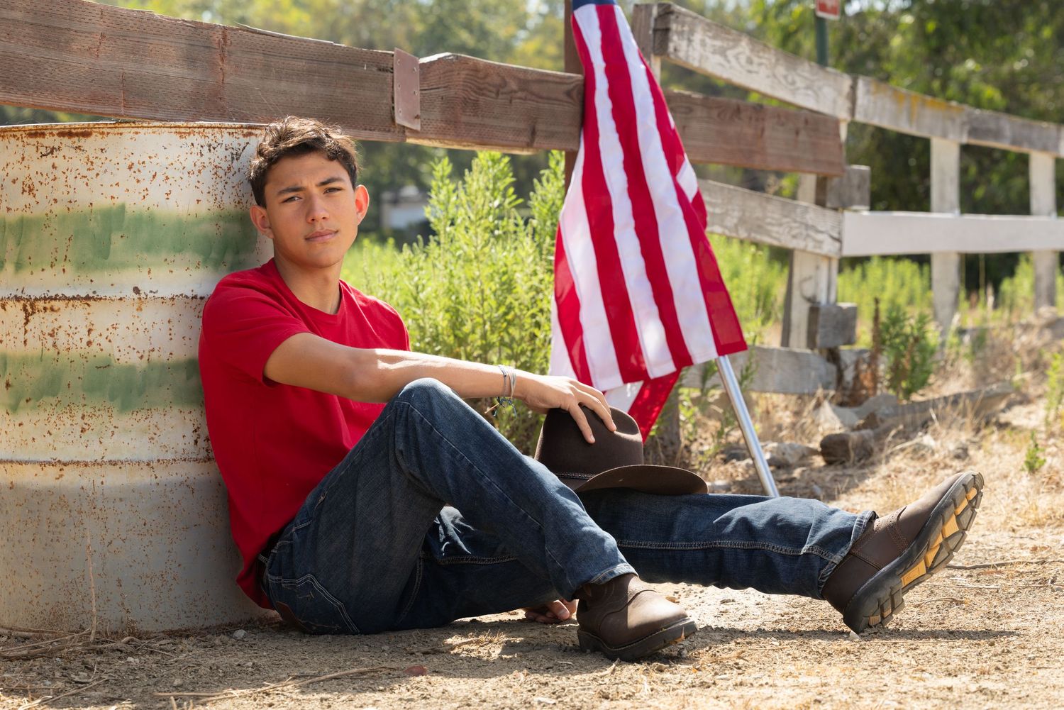 Teen boy in red shirt and jeans sits against fence with American flag outdoors on a sunny day.