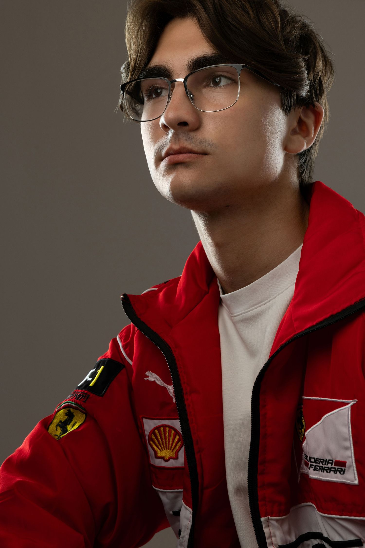 Young man in red Ferrari racing jacket with Shell sponsor patch, wearing glasses, looking upward against gray background.