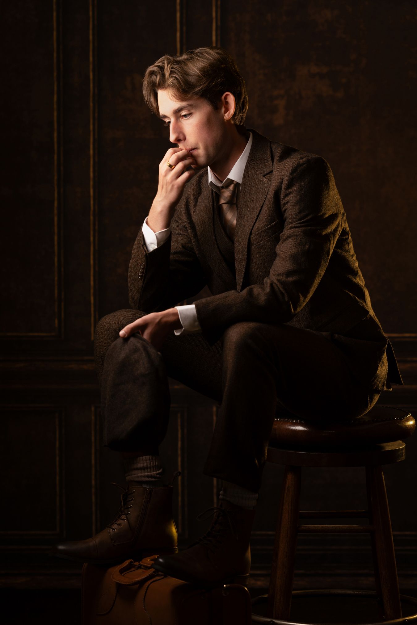 A vintage senior boy in a brown suit sits on a stool in a classic studio, resting his chin on his hand.