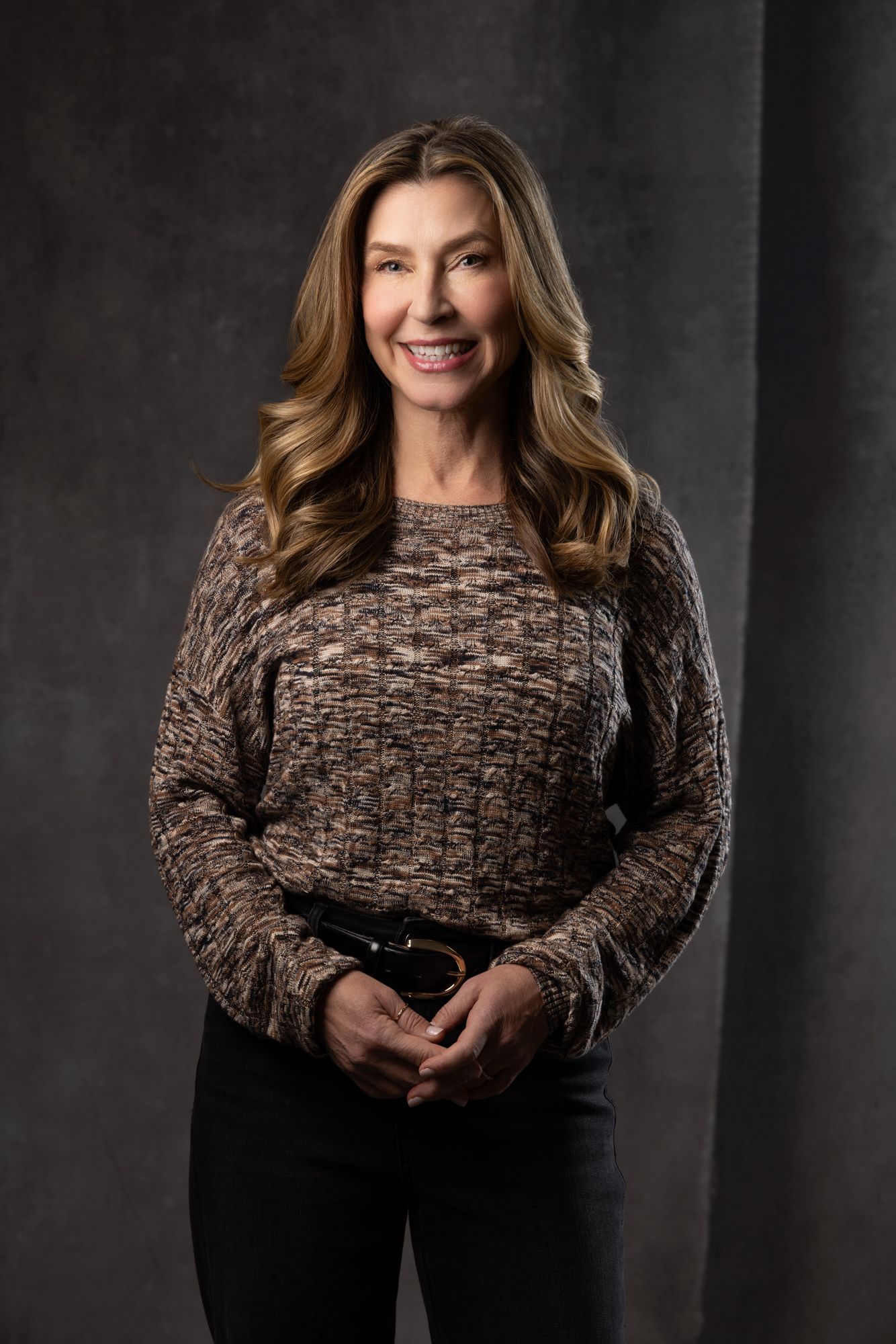 Smiling woman with long brown hair wearing a patterned sweater posing against a gray backdrop.