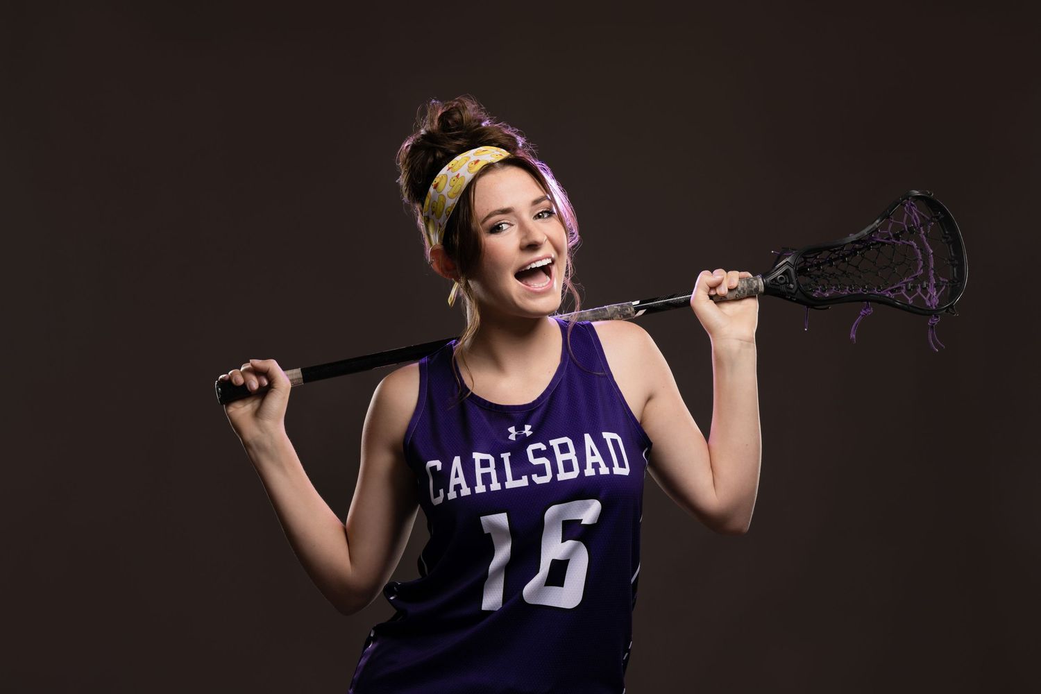 Teen girl in Carlsbad lacrosse jersey number 16 holding stick behind shoulders.