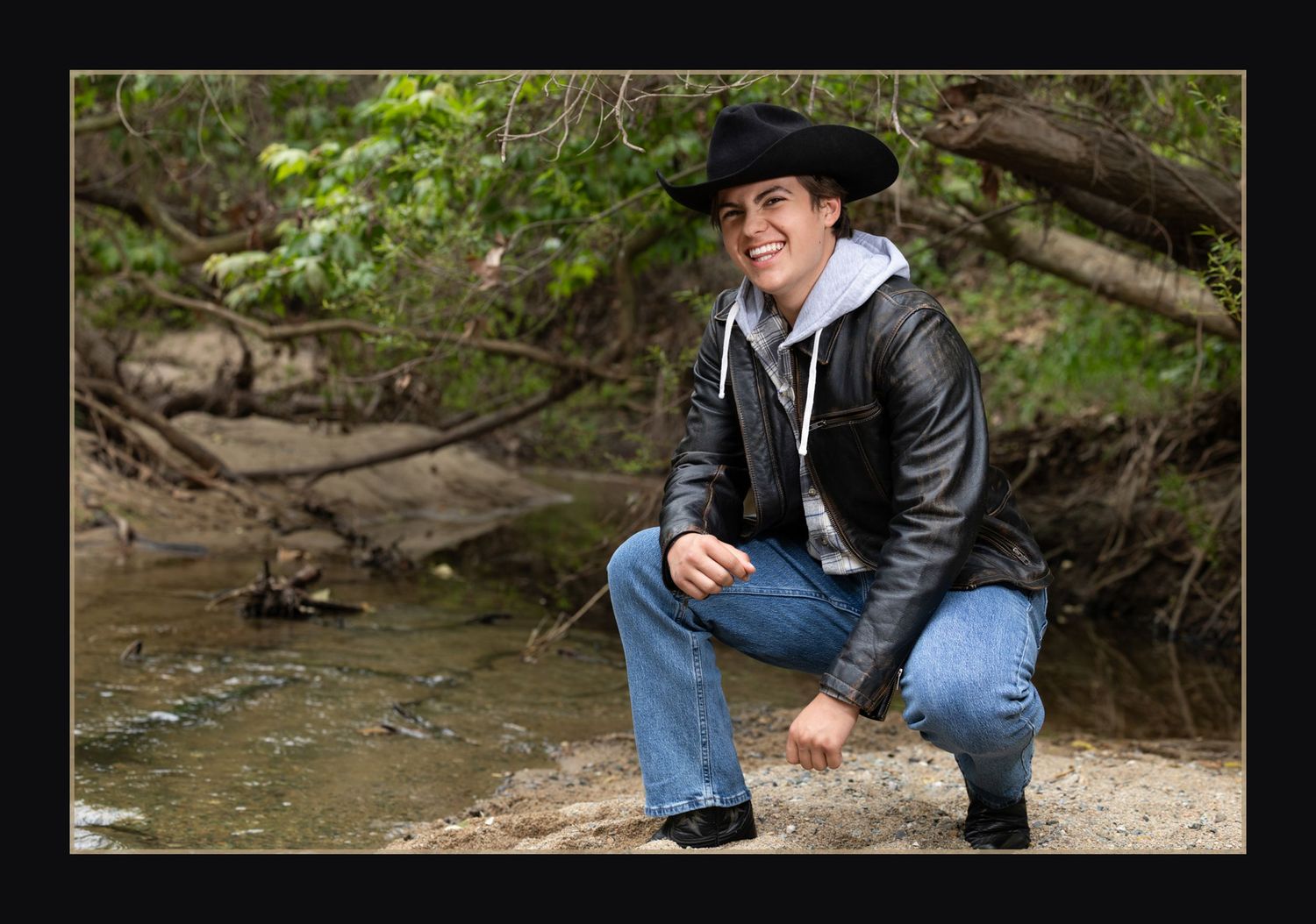 Young man in black cowboy hat and leather jacket crouches by creek in nature.