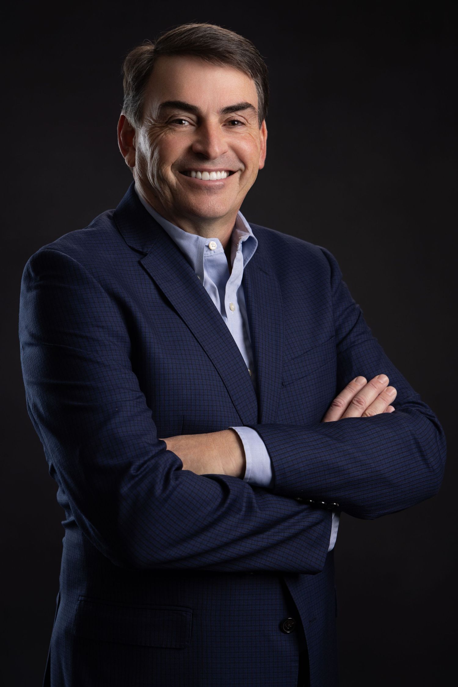 Professional businessman in navy suit smiling with arms crossed against dark background.