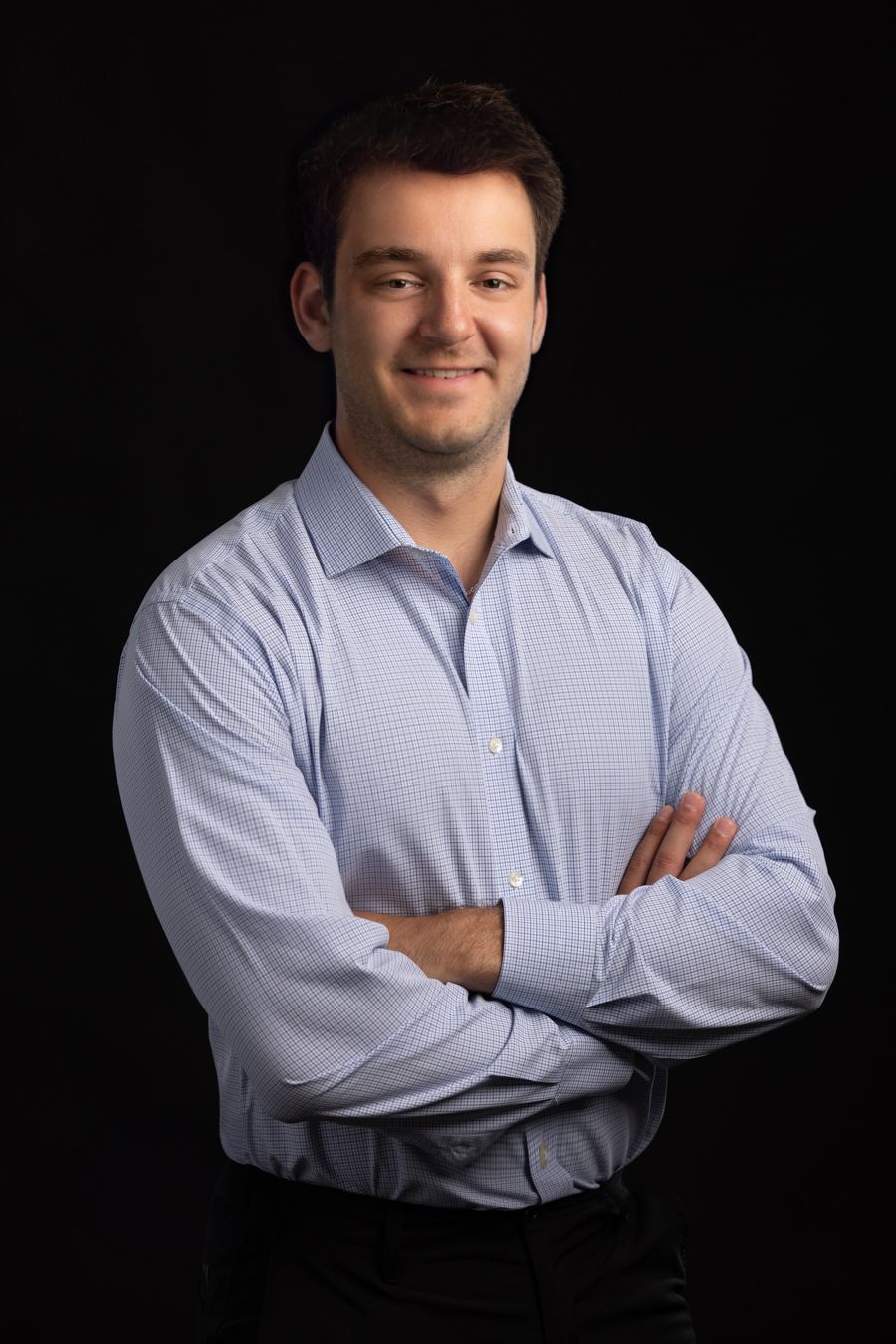 Young professional man in light blue dress shirt smiling with arms crossed against black background.