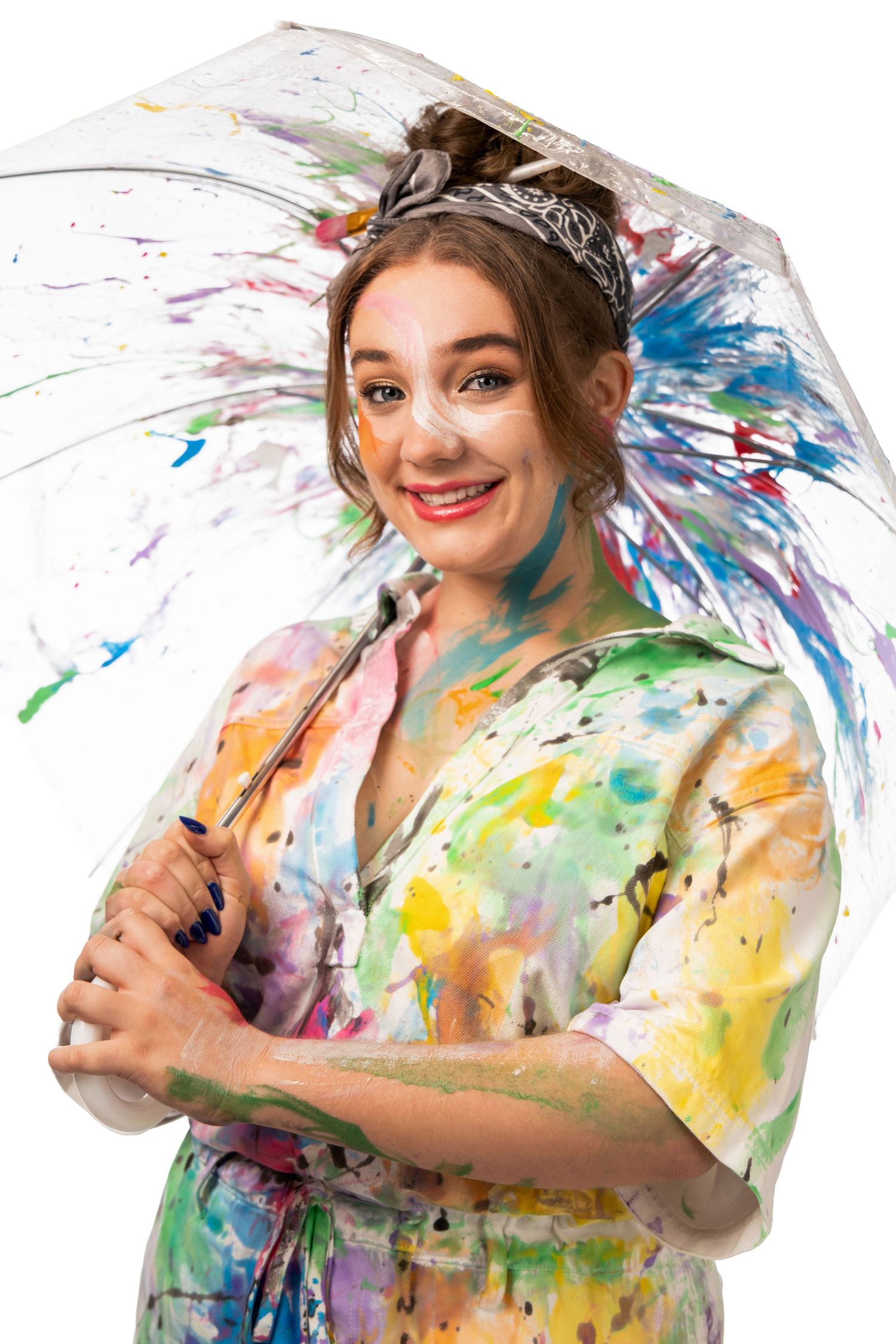 Smiling woman covered in colorful paint splatters holding a transparent umbrella also splattered with paint.