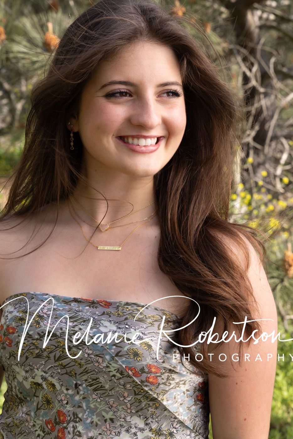 Smiling young woman with long brown hair wearing a floral dress and gold necklace, photographed outdoors by Melanie Robertson Photography.
