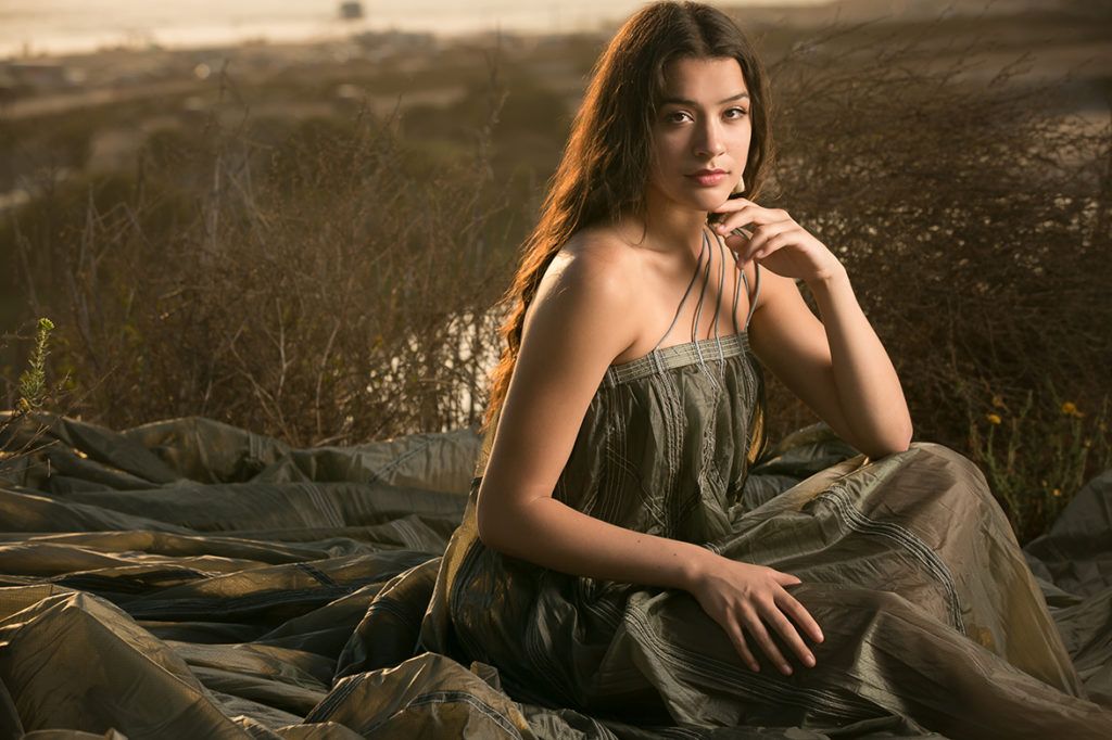 Young woman in olive strapless dress sitting outdoors on fabric in golden hour light.