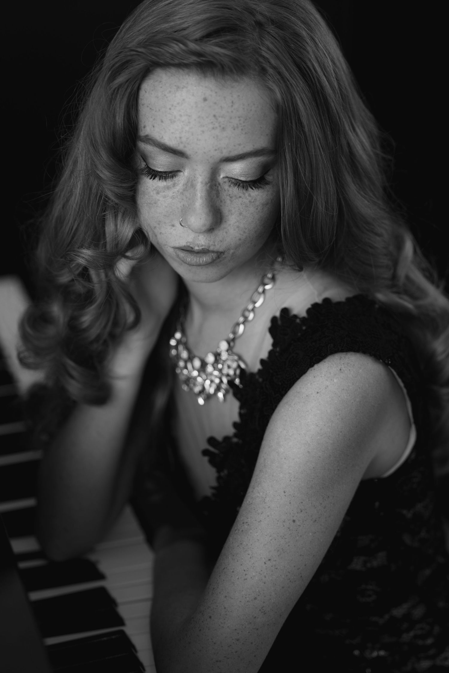 Black and white portrait of a young freckled woman in a black dress and pearl necklace, looking down pensively.
