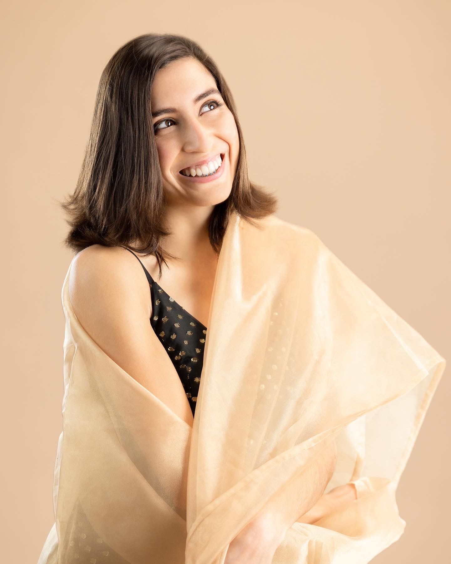 Smiling young woman with dark hair wrapped in a sheer peach scarf over a black top against a beige background.