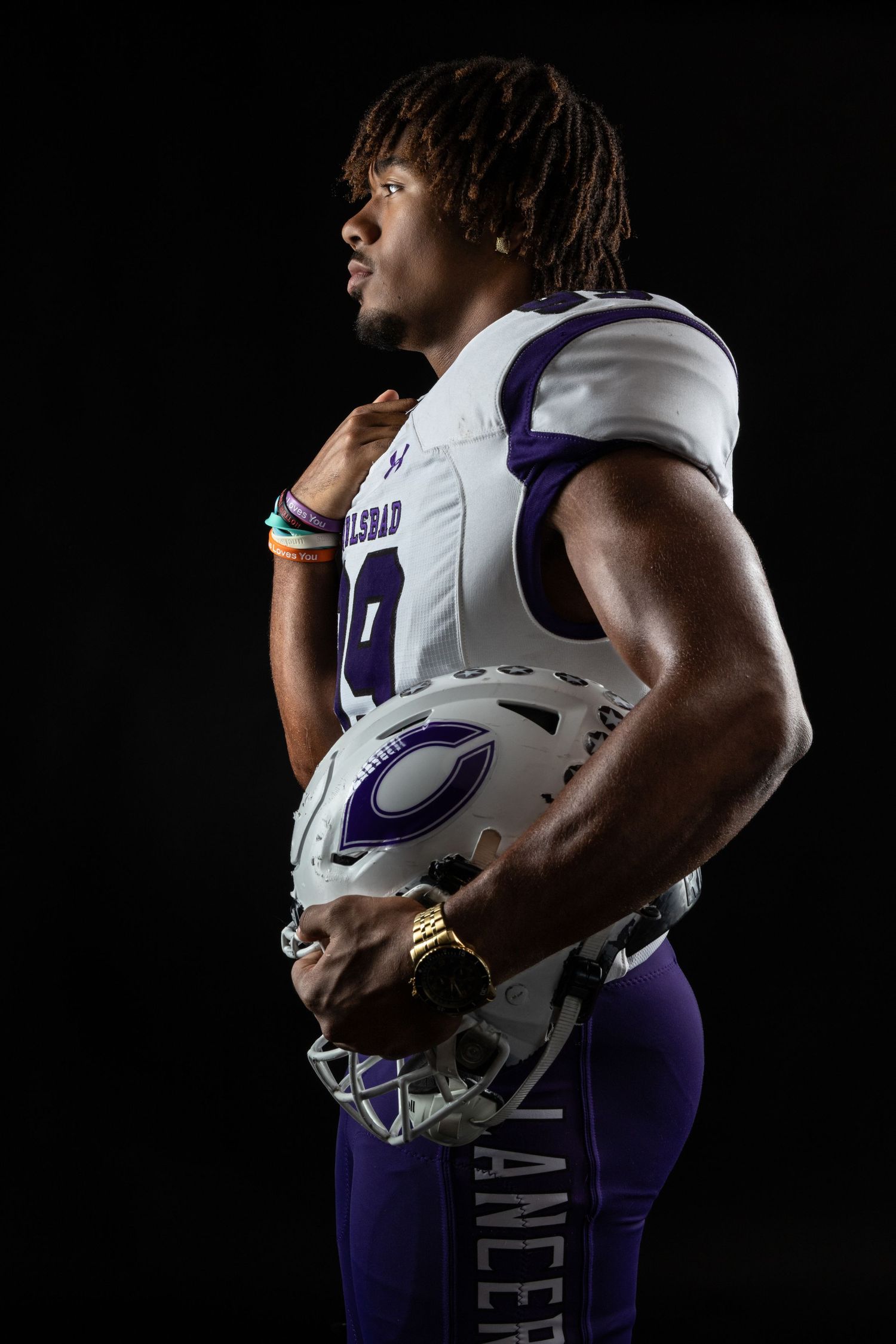 Football player in white and purple uniform holds helmet against black background.